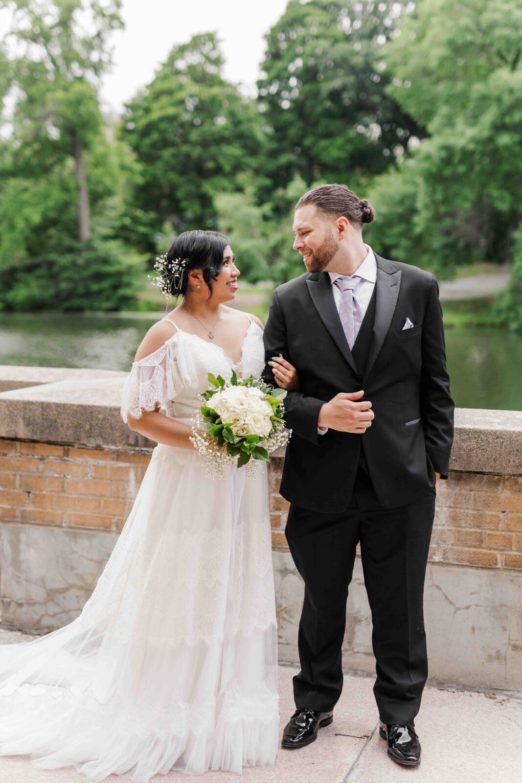Bride in a white dress holding a bouquet stands arm-in-arm with a groom in a black suit outdoors by a stone railing and greenery.