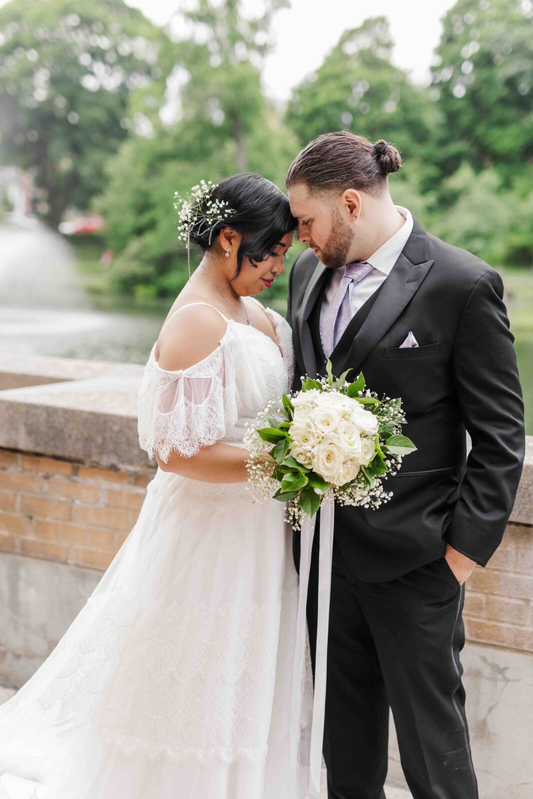 Bride and groom stand close together outdoors, touching foreheads. The bride holds a bouquet of white roses and wears a white gown; the groom wears a black suit with a light tie.