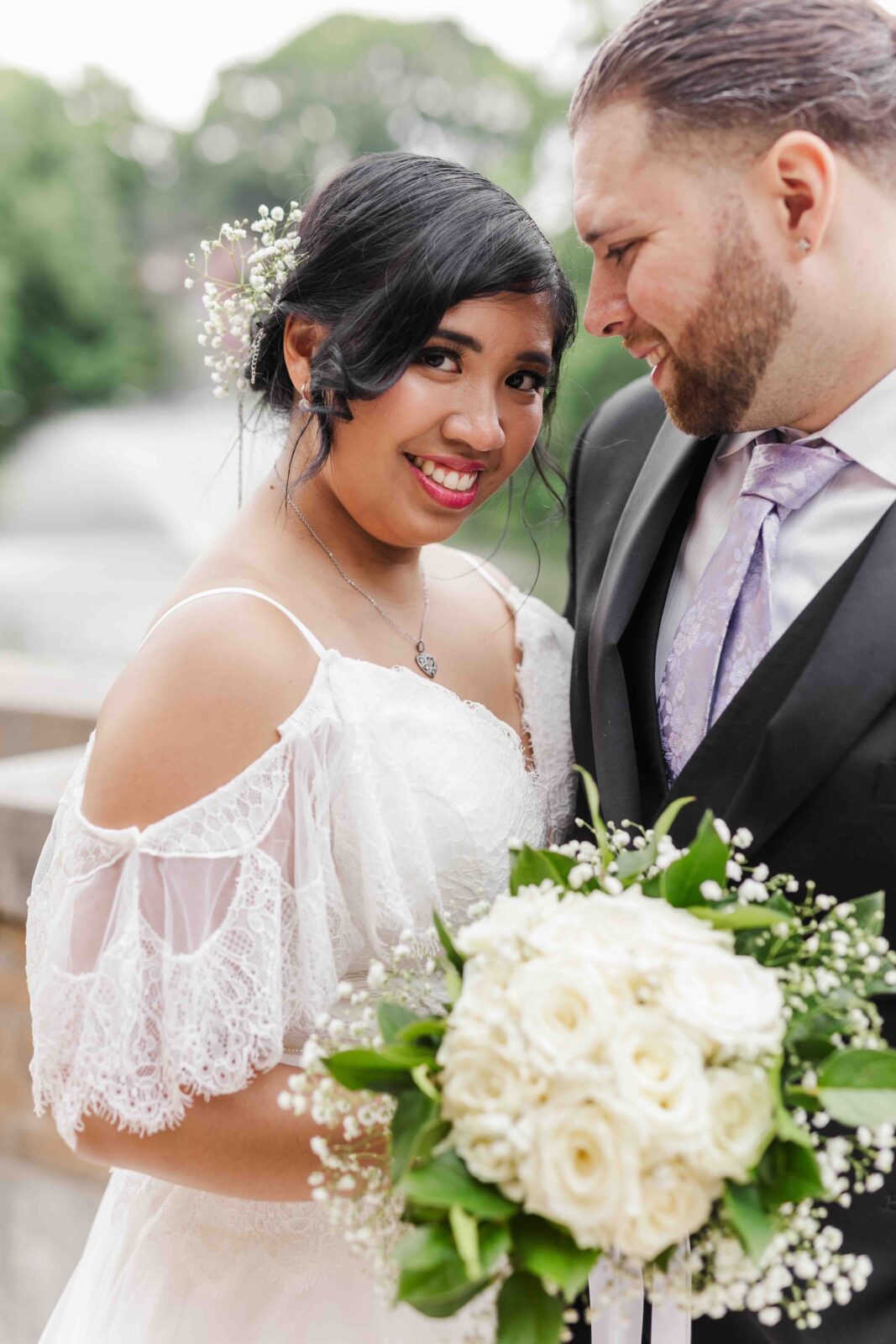 Bride in a white lace dress holding a bouquet of white roses stands next to groom in a suit outdoors, both smiling.