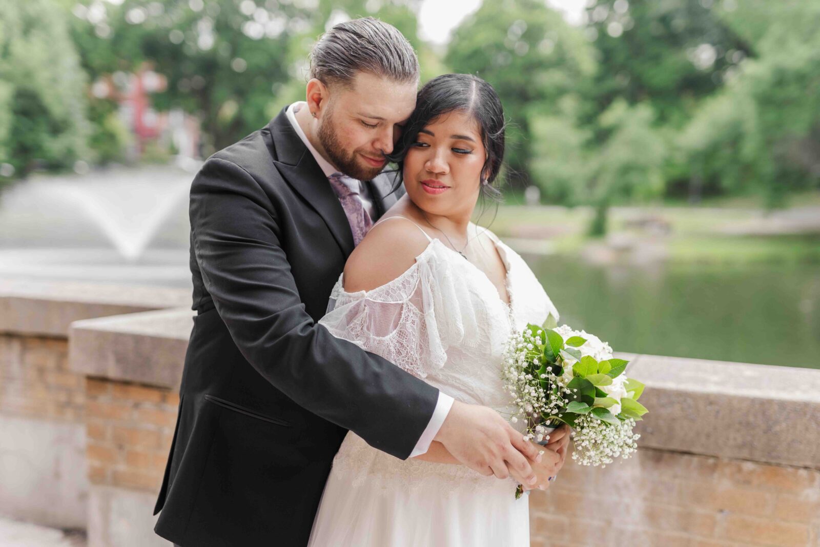 A bride and groom stand close together outdoors by a stone railing and pond. The groom embraces the bride from behind as she holds a bouquet of white flowers and greenery.