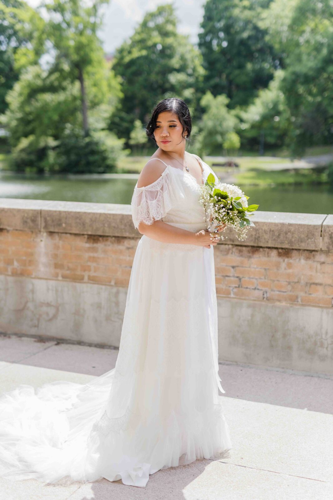 A woman in a white off-shoulder wedding dress holds a bouquet and stands outdoors by a brick wall with greenery and water in the background.