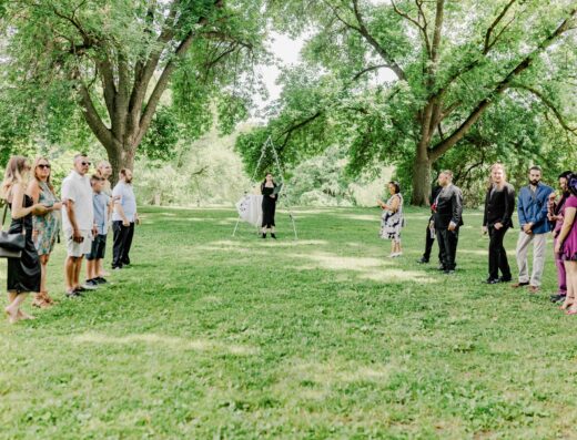 A group of people stands in two lines outdoors on a grassy area, facing each other with trees in the background and an officiant at the center.