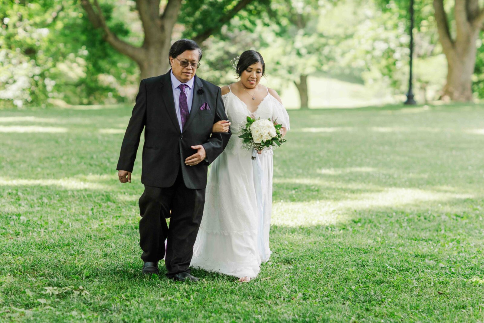 A woman in a white dress holding a bouquet walks arm in arm with a man in a dark suit across a grassy area with trees in the background.
