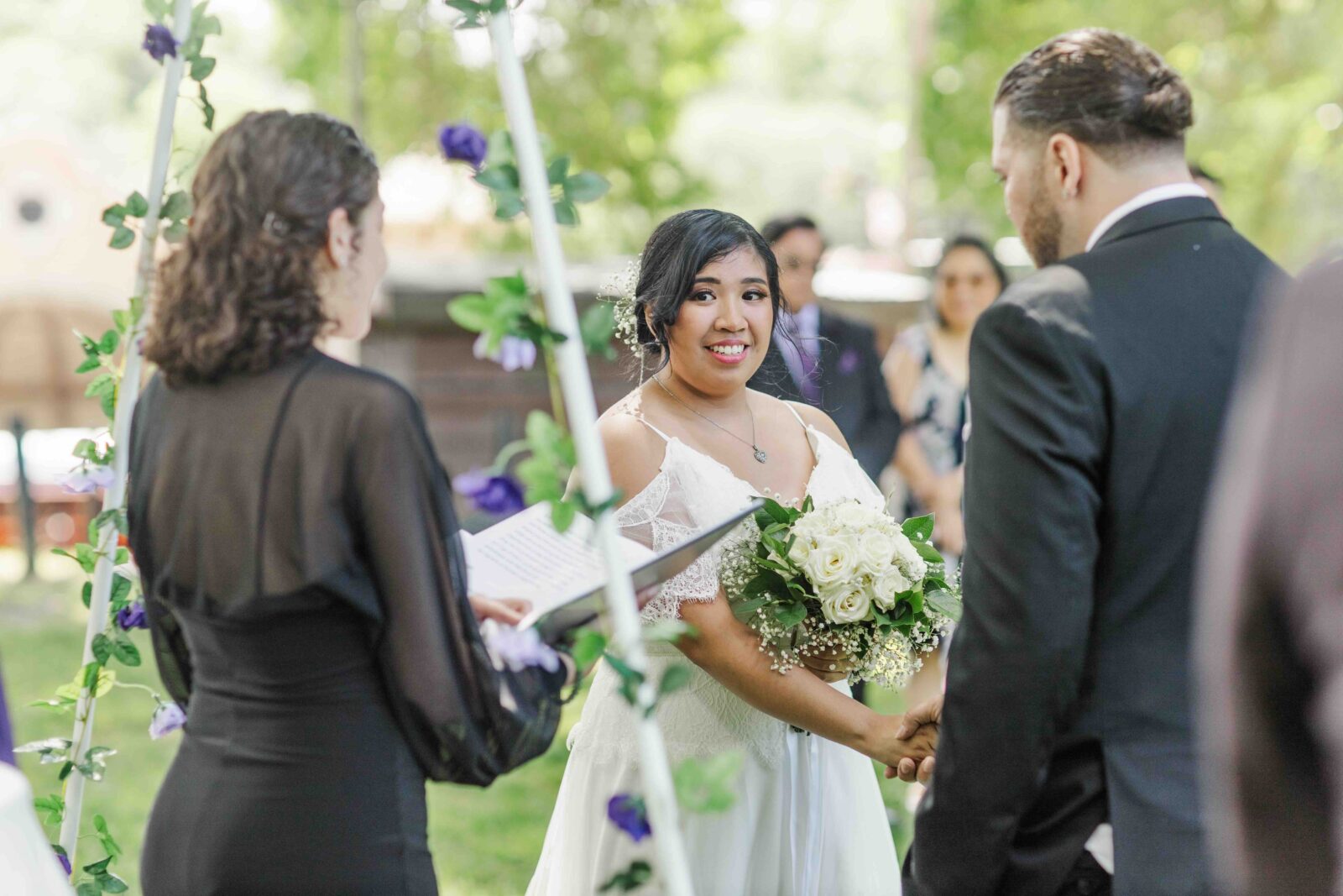 Bride and groom stand facing each other and holding hands during an outdoor wedding ceremony, with an officiant reading from a book.