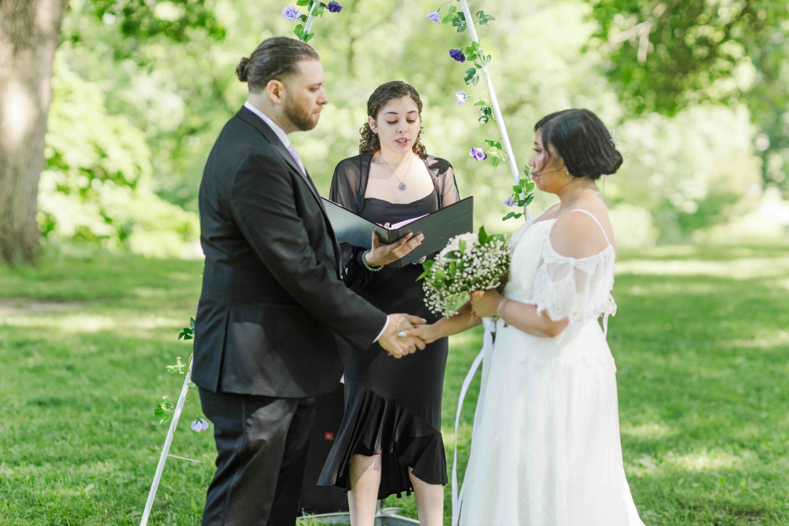 A couple stands facing each other holding hands under a floral arch while an officiant reads from a book outdoors.