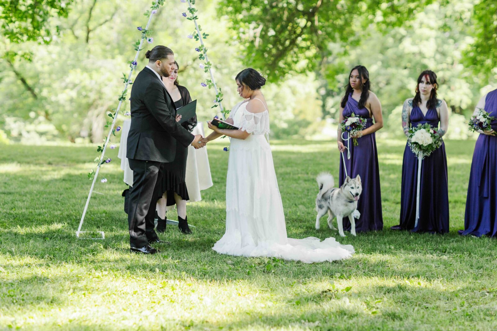 A couple stands facing each other during an outdoor wedding ceremony, accompanied by bridesmaids in purple dresses and a dog on a leash.