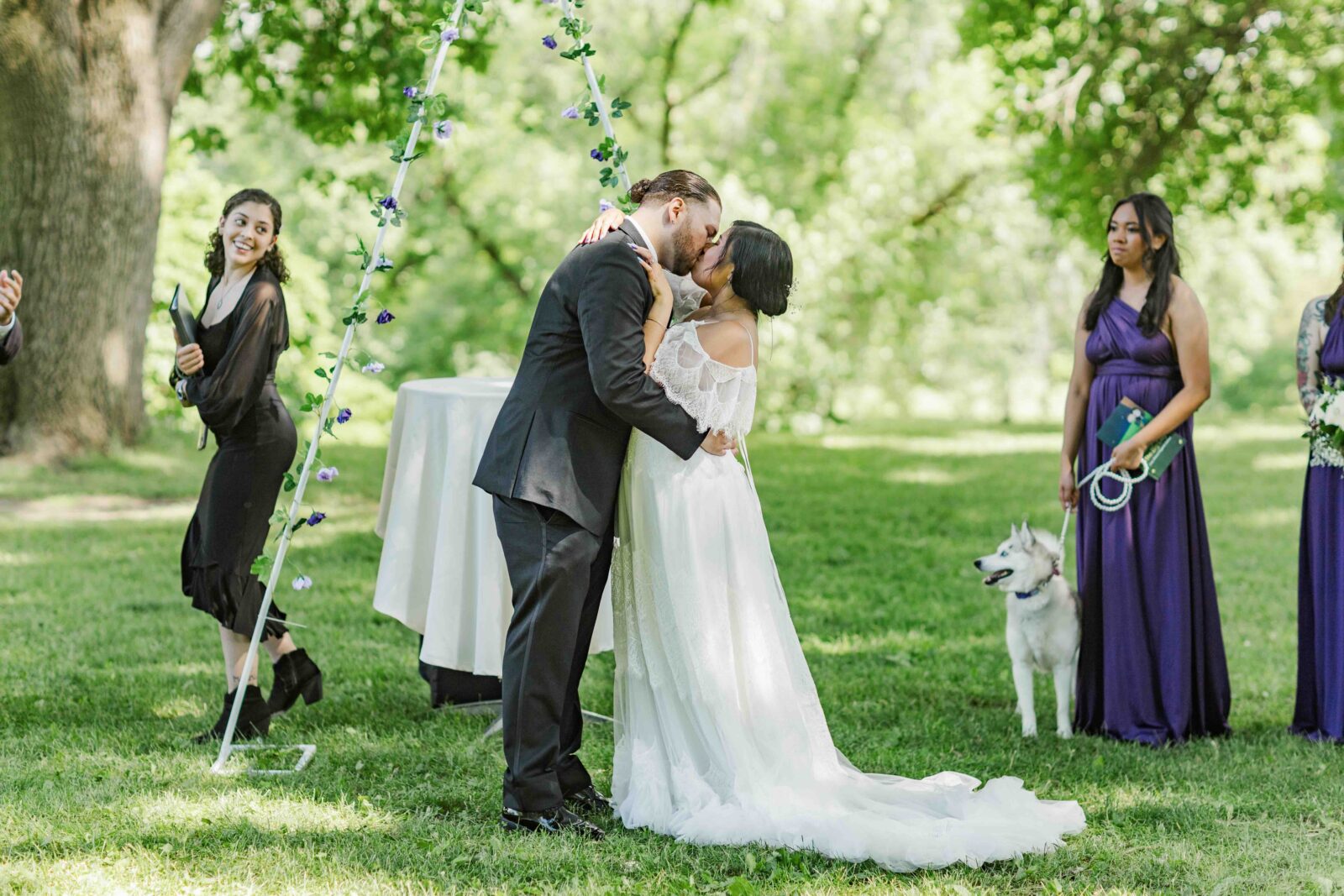 A bride and groom kiss at an outdoor wedding ceremony, surrounded by bridesmaids and a dog, with trees and greenery in the background.