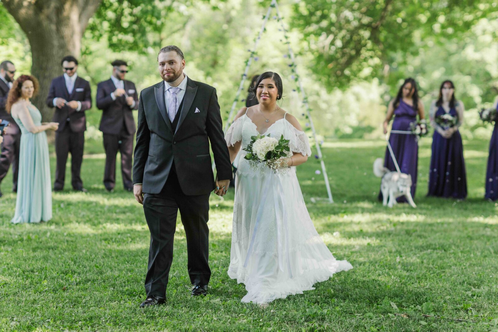 A bride in a white dress and a groom in a black suit walk together outdoors, holding hands, with people and trees in the background.