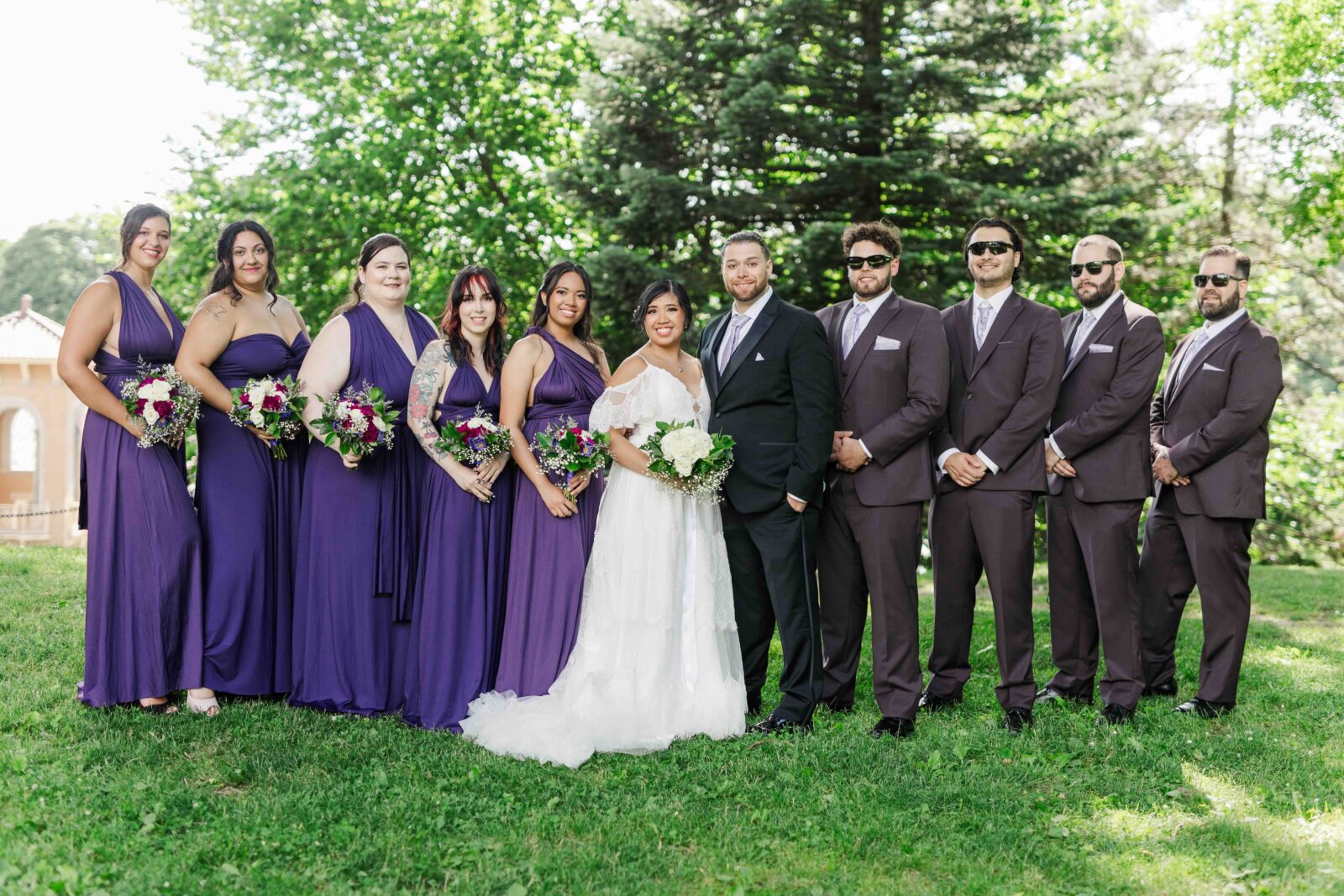A bride and groom stand outdoors with their wedding party; bridesmaids wear purple dresses, groomsmen wear dark suits, and trees are visible in the background.