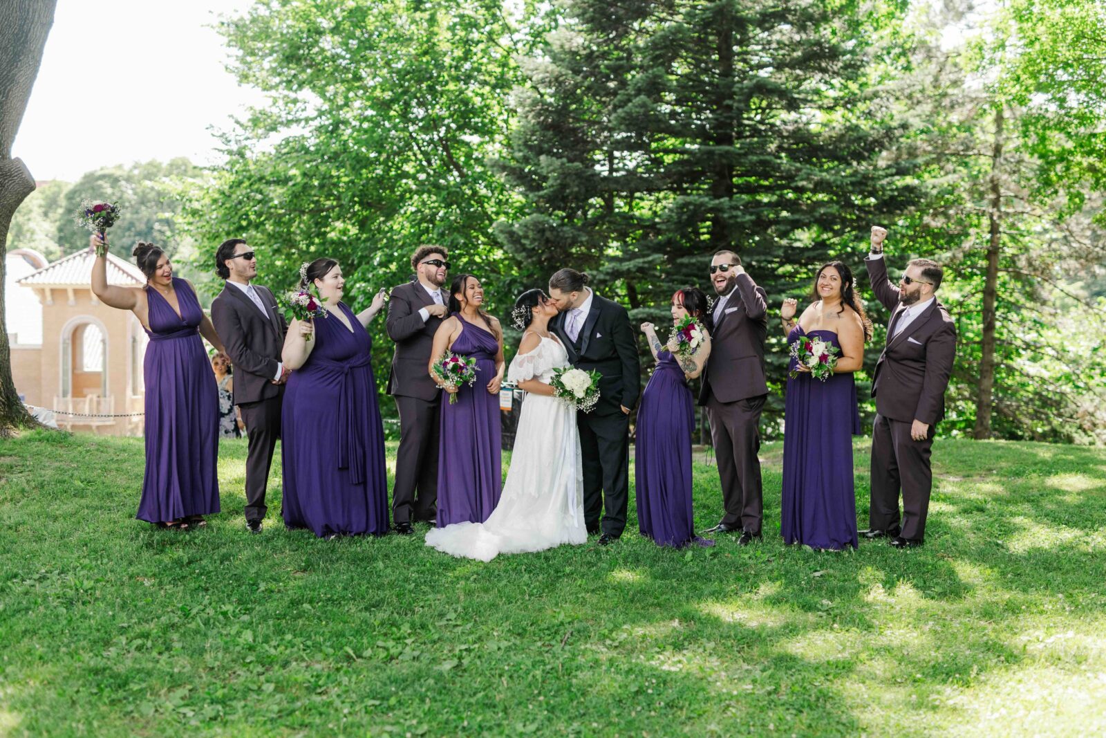 A bride and groom kiss outdoors, surrounded by their wedding party dressed in purple gowns and dark suits, with trees and greenery in the background.