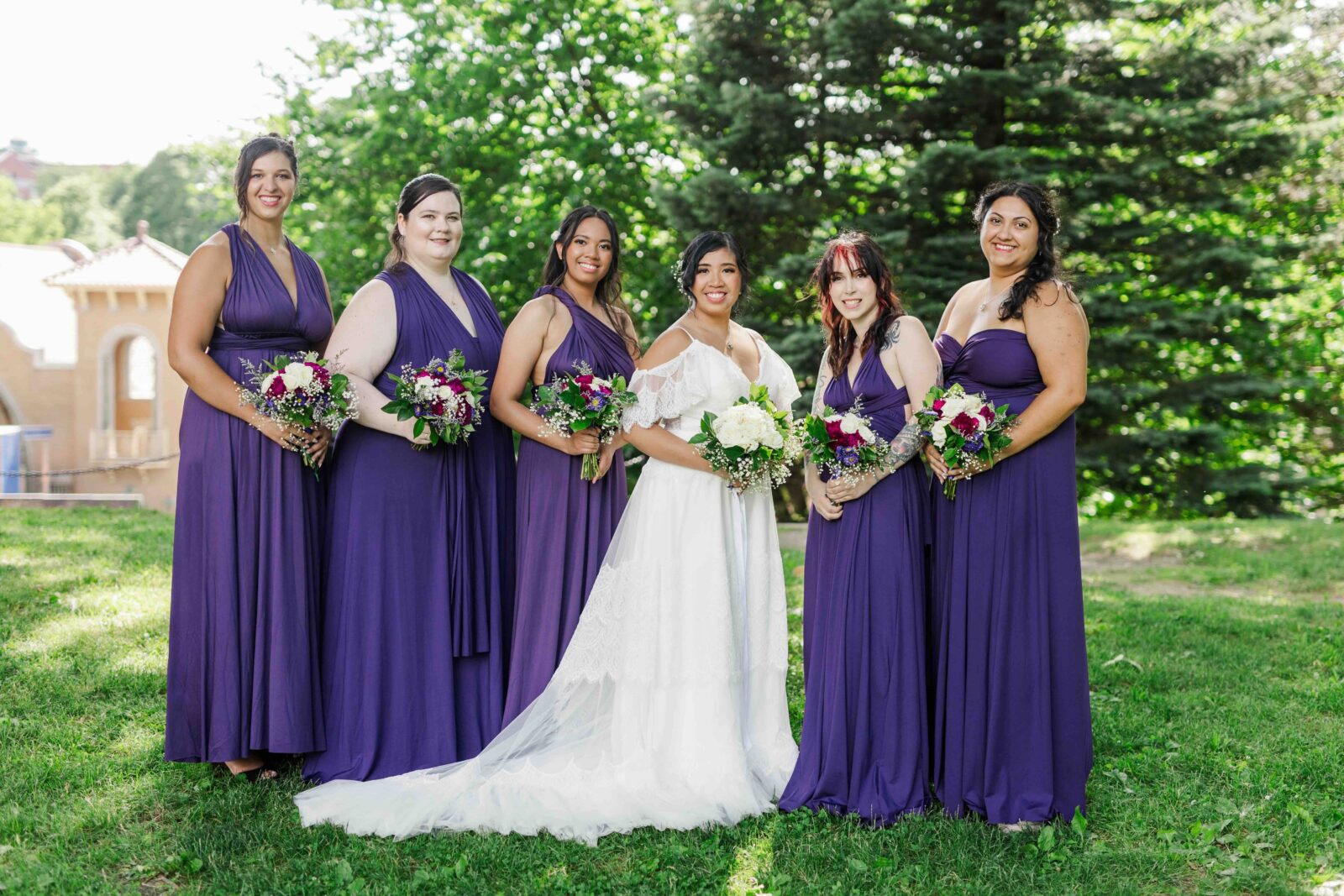 Five bridesmaids in deep royal purple convertible floor-length gowns with mixed necklines hold white and magenta bouquets beside the bride outdoors at Washington Park in Albany