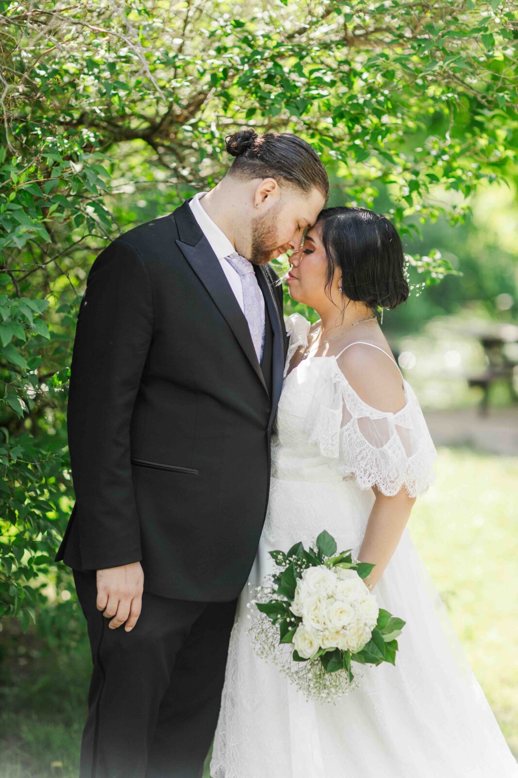 A bride and groom stand close together outdoors, touching foreheads. The bride holds a bouquet of white flowers and wears a white dress; the groom is in a black suit. Greenery surrounds them.