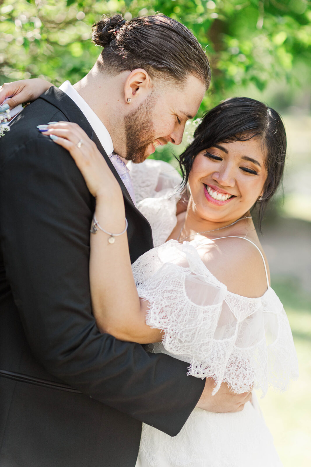 A bride and groom in wedding attire embrace outdoors, both smiling with eyes closed, surrounded by greenery and sunlight.