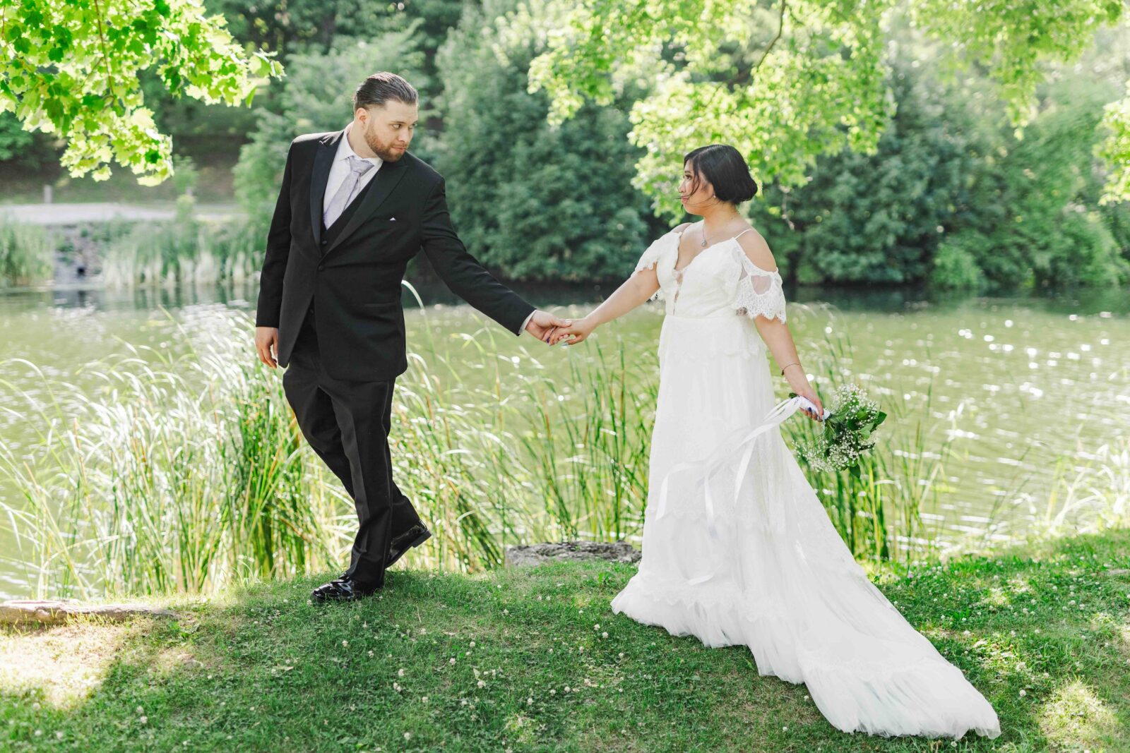 A bride and groom hold hands by a lakeside, surrounded by greenery. The bride wears a white dress and holds a bouquet, while the groom wears a black suit.