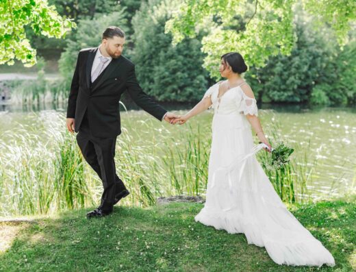 A bride and groom hold hands by a lakeside, surrounded by greenery. The bride wears a white dress and holds a bouquet, while the groom wears a black suit.