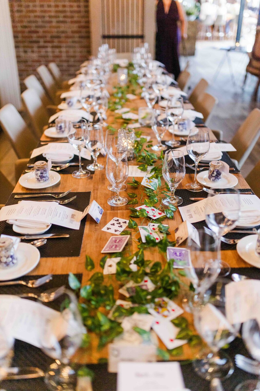 A long wooden table set for dinner, decorated with green leaves, playing cards, glassware, menus, and small gift boxes.