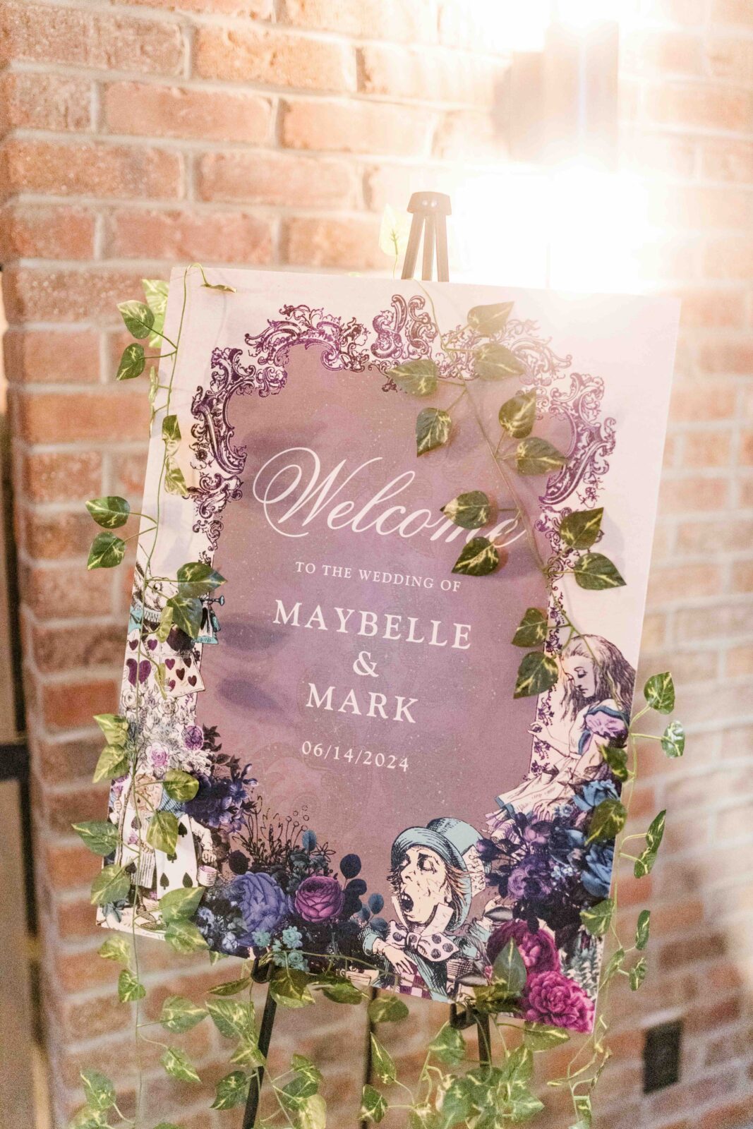 Wedding sign reads “Welcome to the wedding of Maybelle & Mark 06/14/2024,” decorated with vines and Alice in Wonderland illustrations, set against a brick wall.