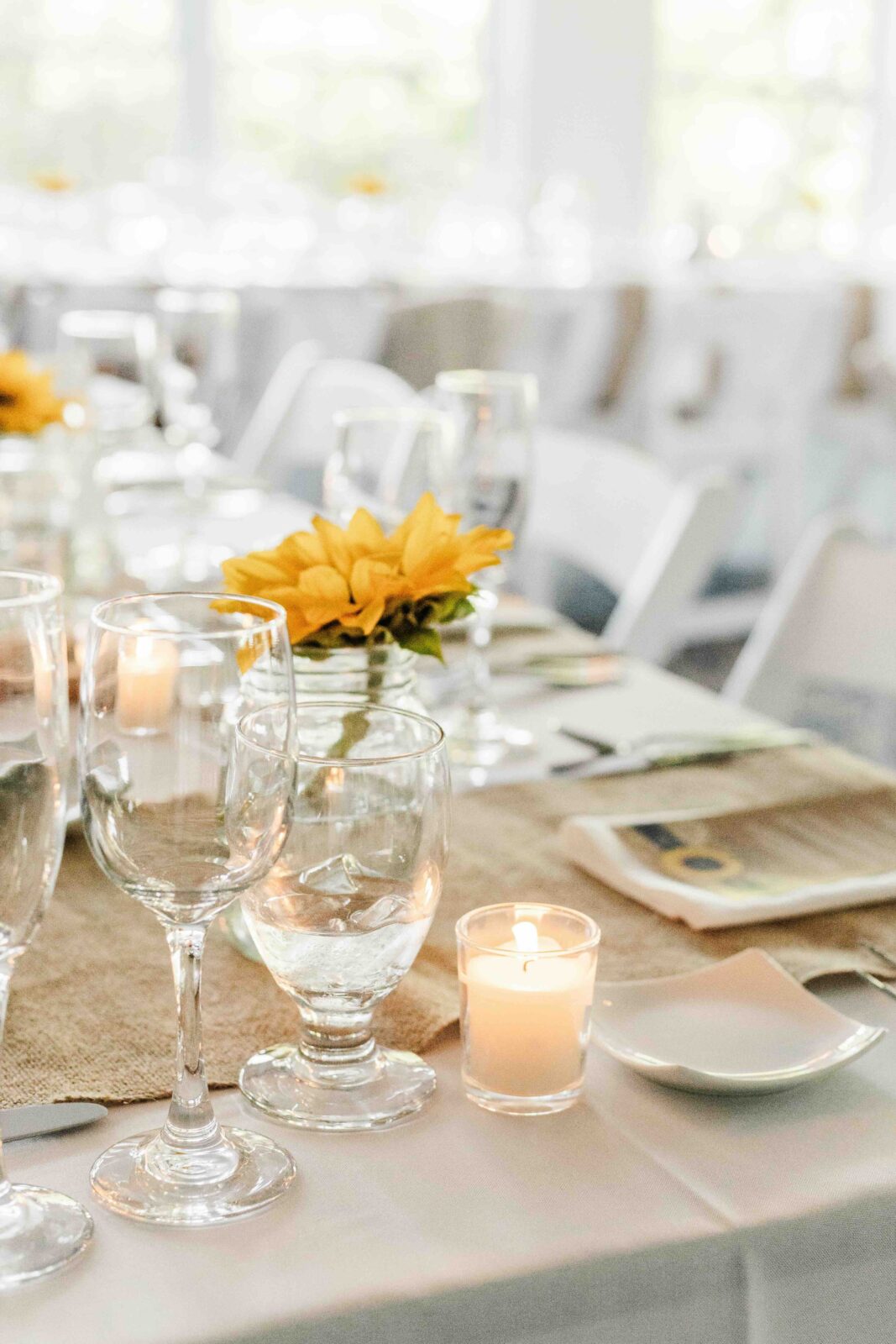 Table set with glassware, a lit votive candle, white plates, and a sunflower centerpiece on a burlap runner in a bright, airy room.