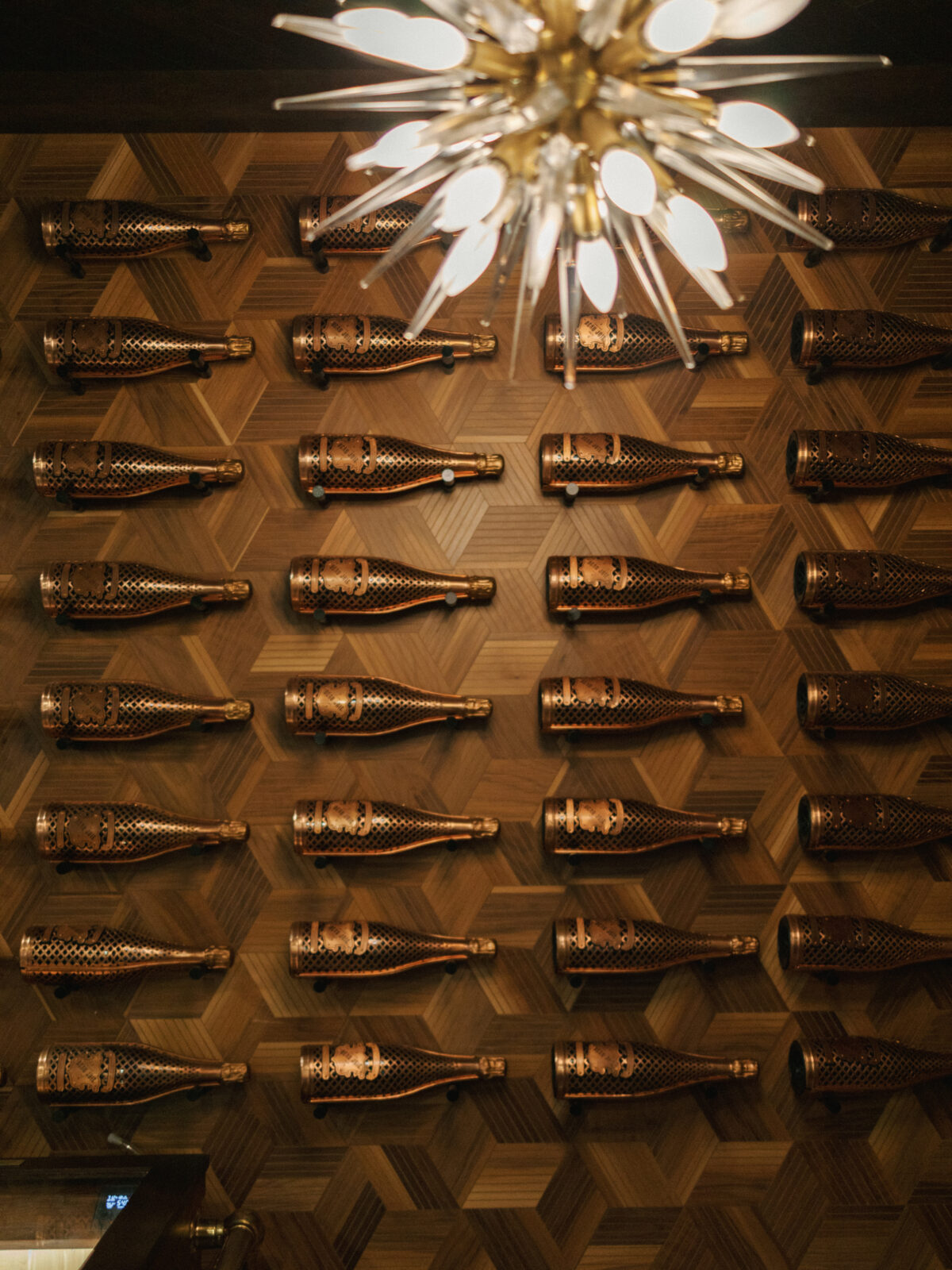 Metal wine bottle holders mounted in a grid pattern on a geometric wooden wall beneath a modern chandelier.