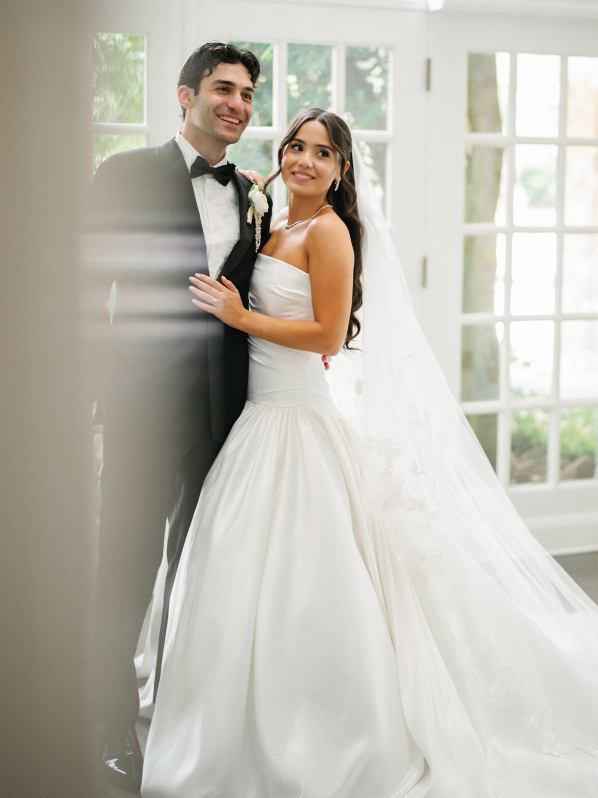 Bride in a white gown and groom in a black tuxedo stand together indoors, smiling, with natural light and greenery visible through glass doors behind them.