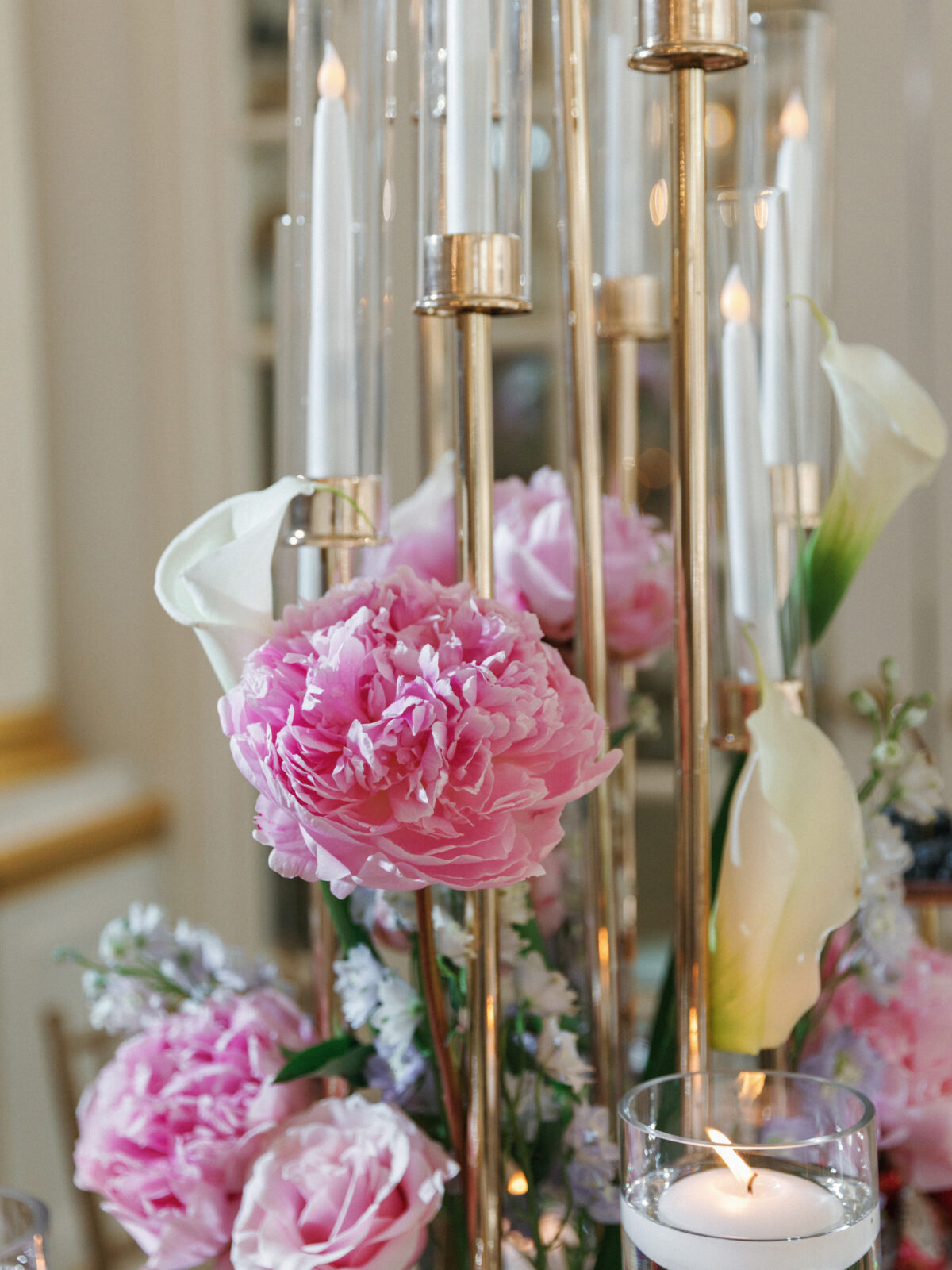 Pink peonies, white calla lilies, and candles in glass holders arranged together on a gold centerpiece stand.