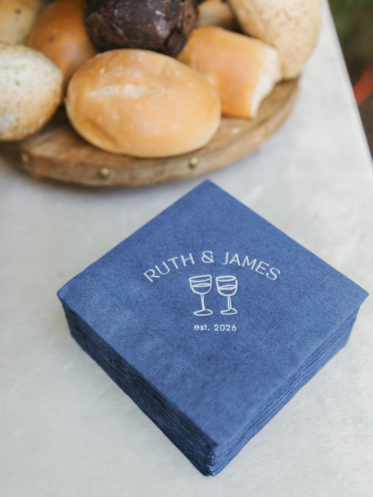 A stack of blue napkins with “Ruth & James est. 2026” and two wine glasses printed on them sits beside a wooden tray with assorted bread rolls.