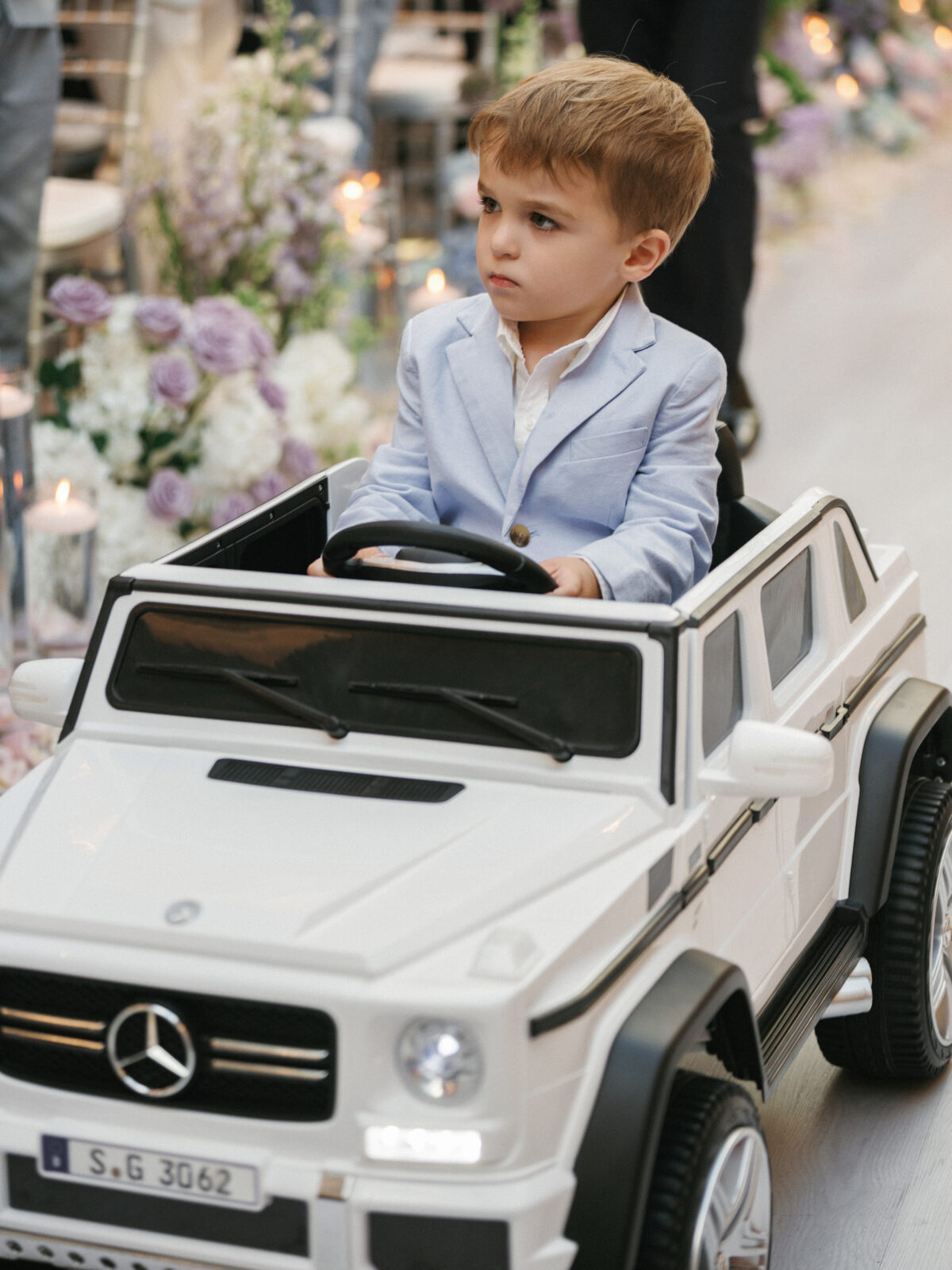 Young boy in a light blue blazer sits in a white toy Mercedes car, surrounded by flowers and candles indoors.