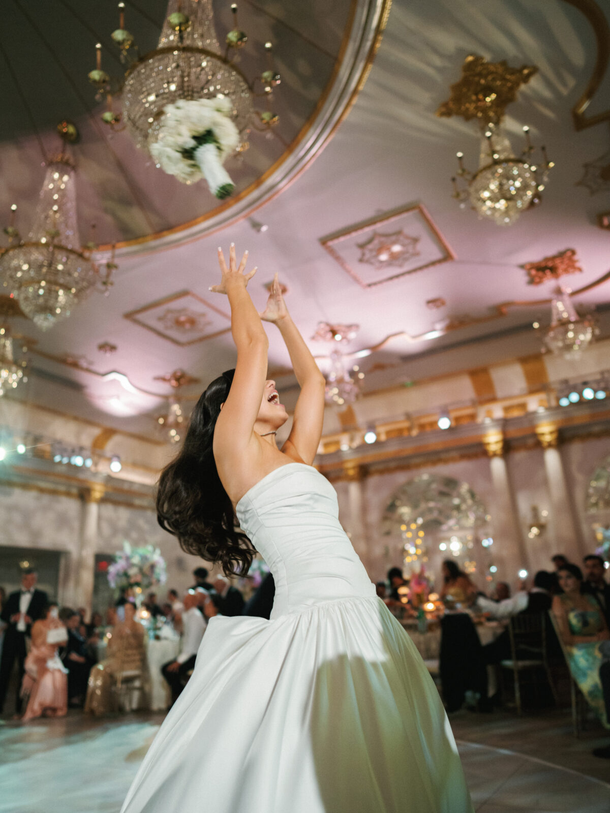 A bride in a white gown throws her bouquet into the air during a wedding reception in a grand, chandelier-lit ballroom.