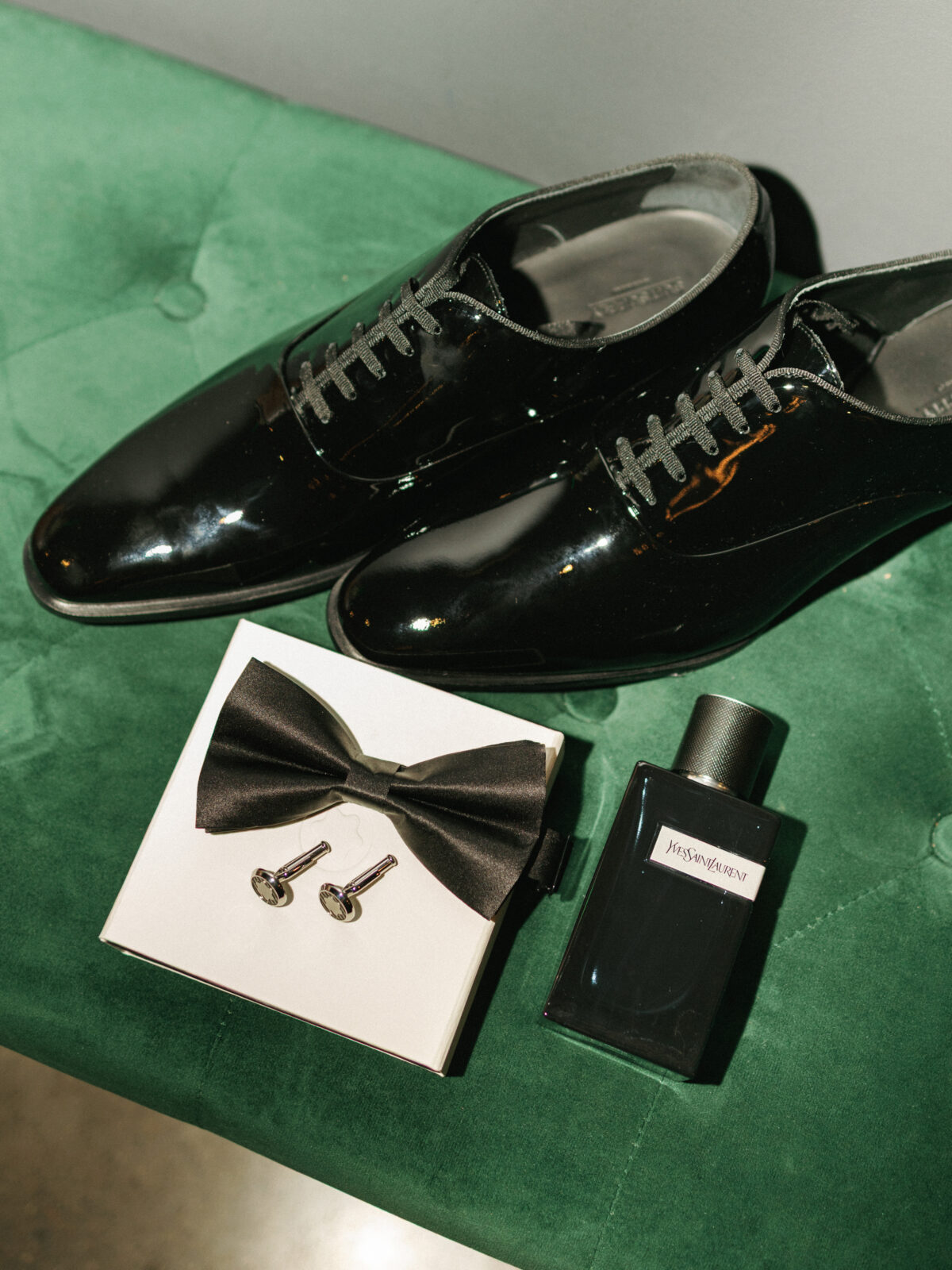 Black dress shoes, a black bow tie, silver cufflinks, and a bottle of Yves Saint Laurent cologne arranged on a green velvet surface.