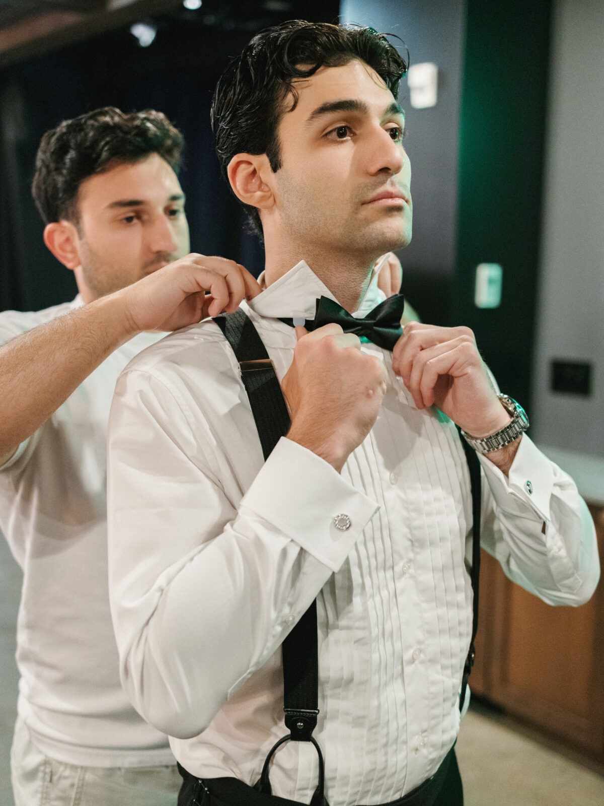 A man in formal attire adjusts his bow tie while another man stands behind him, helping fix his collar.