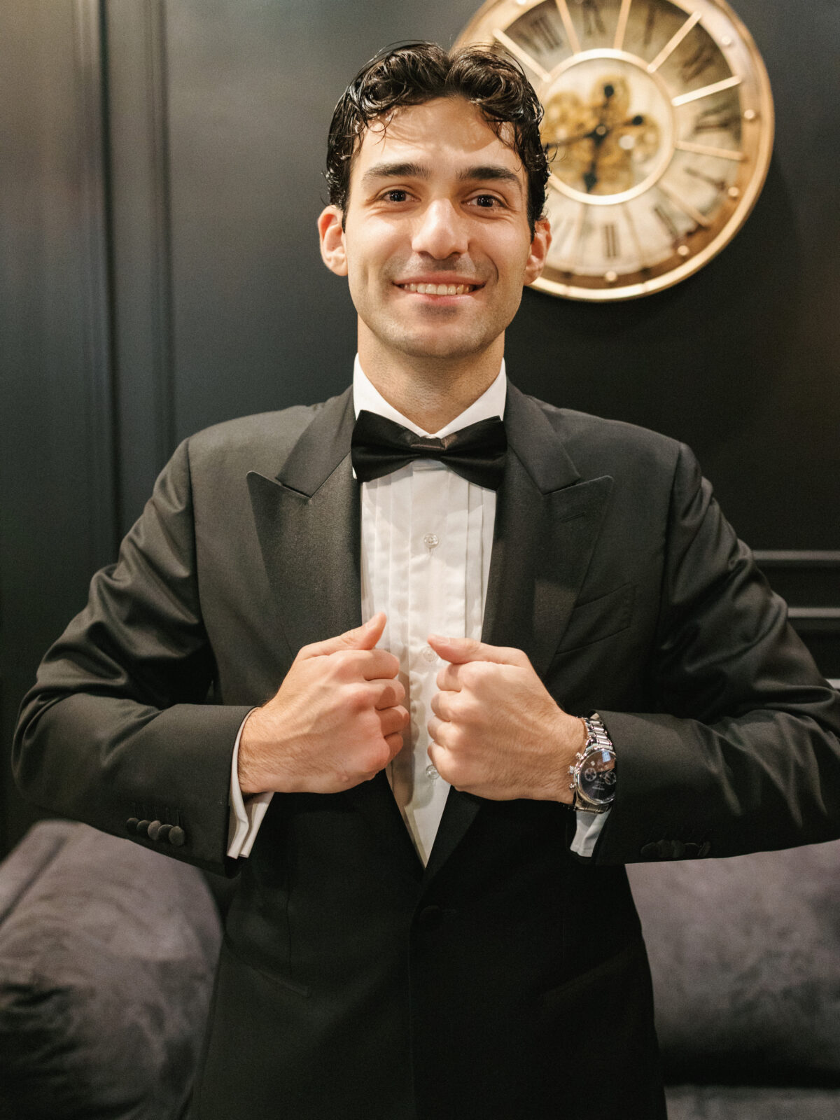 A man in a black tuxedo adjusts his jacket and smiles at the camera. A large clock is visible on the dark wall behind him.