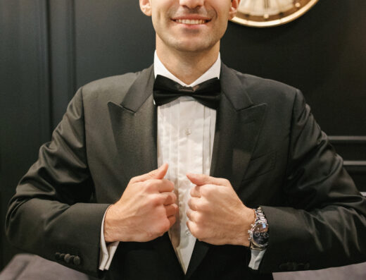 A man in a black tuxedo adjusts his jacket and smiles at the camera. A large clock is visible on the dark wall behind him.