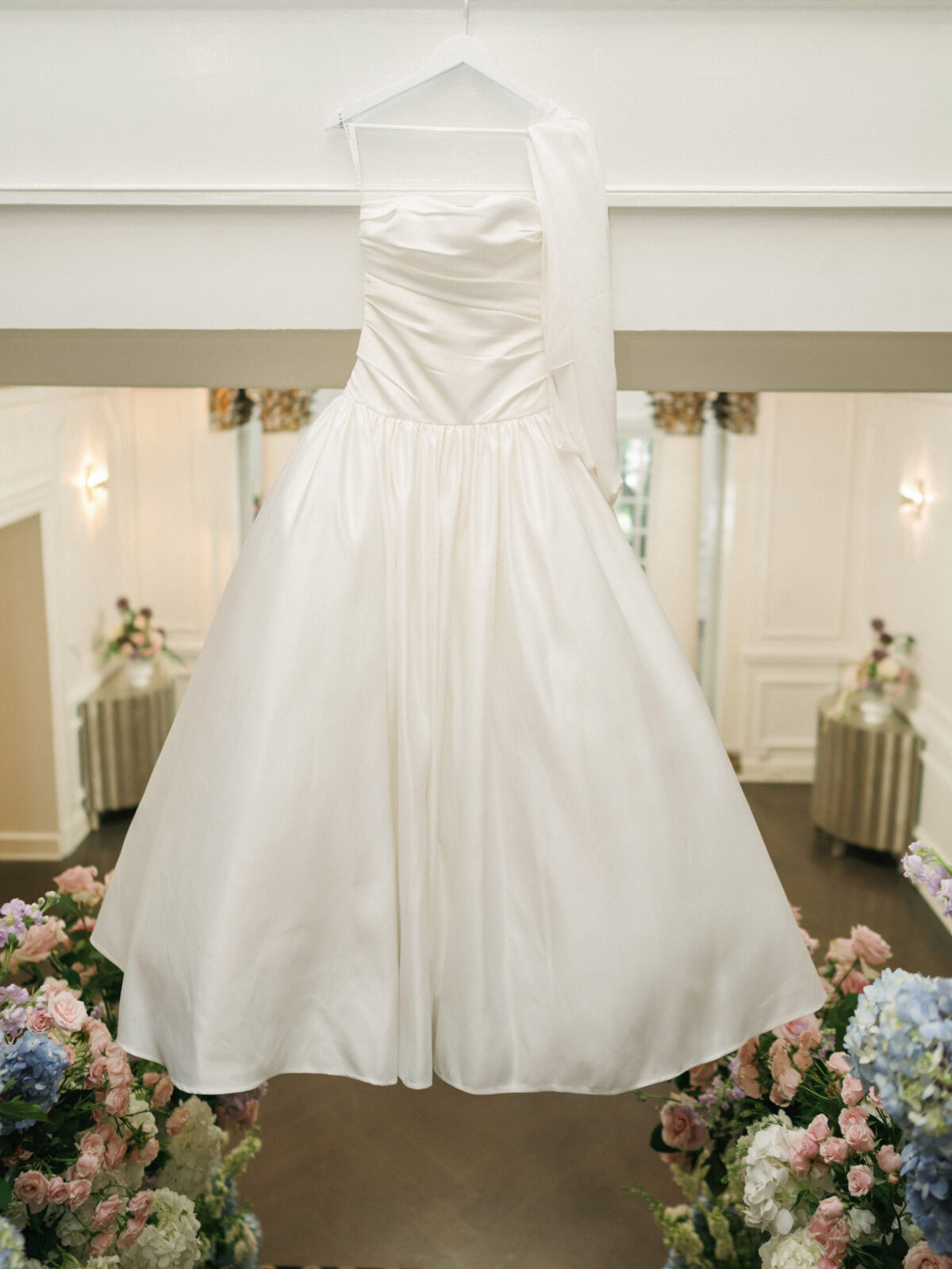 A white satin wedding dress with a full skirt hangs on a hanger in a bright, elegant room decorated with pastel flowers.