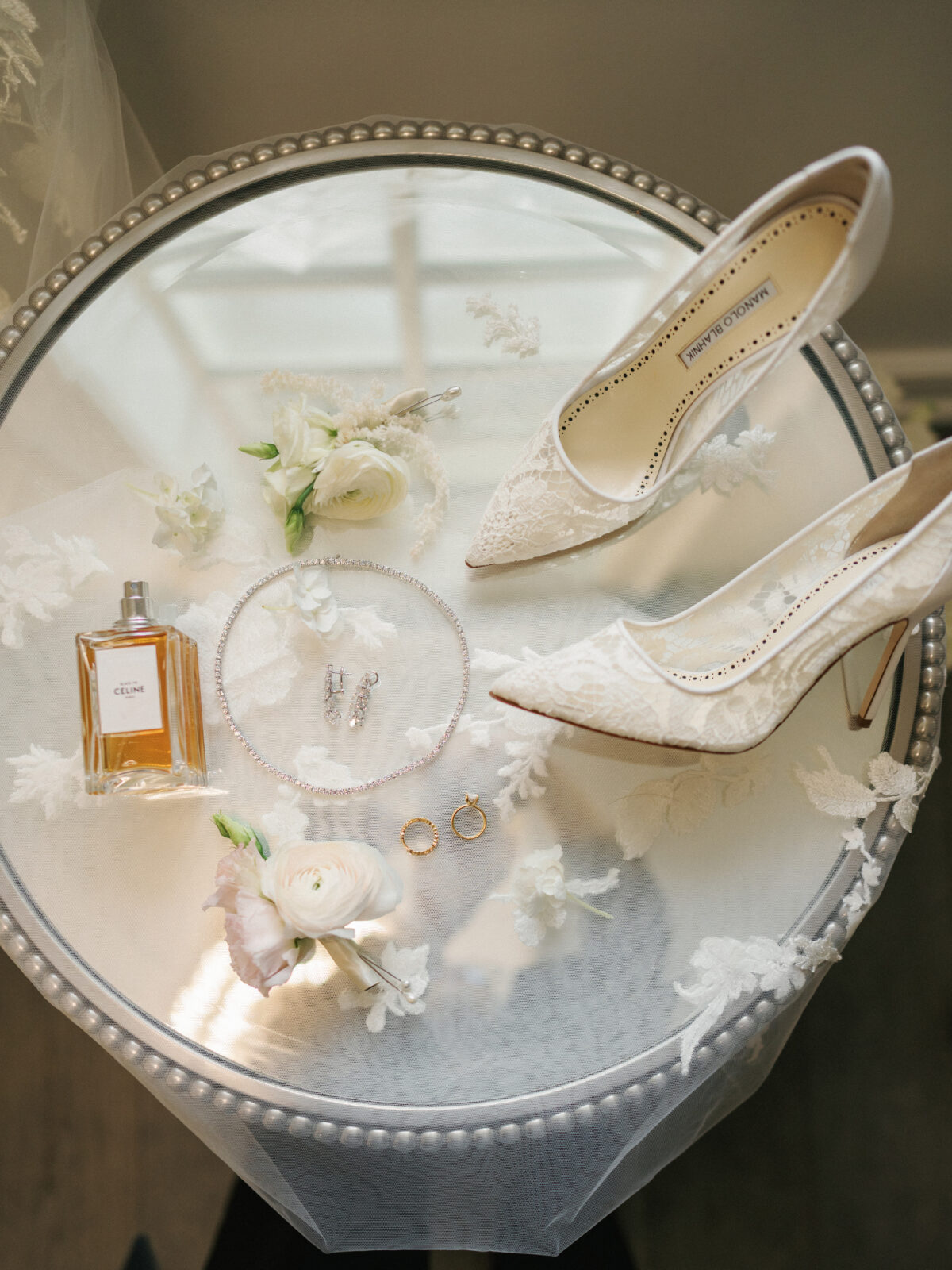 A round tray with white lace heels, a perfume bottle, a necklace, earrings, two rings, and white floral arrangements neatly arranged on a reflective surface.