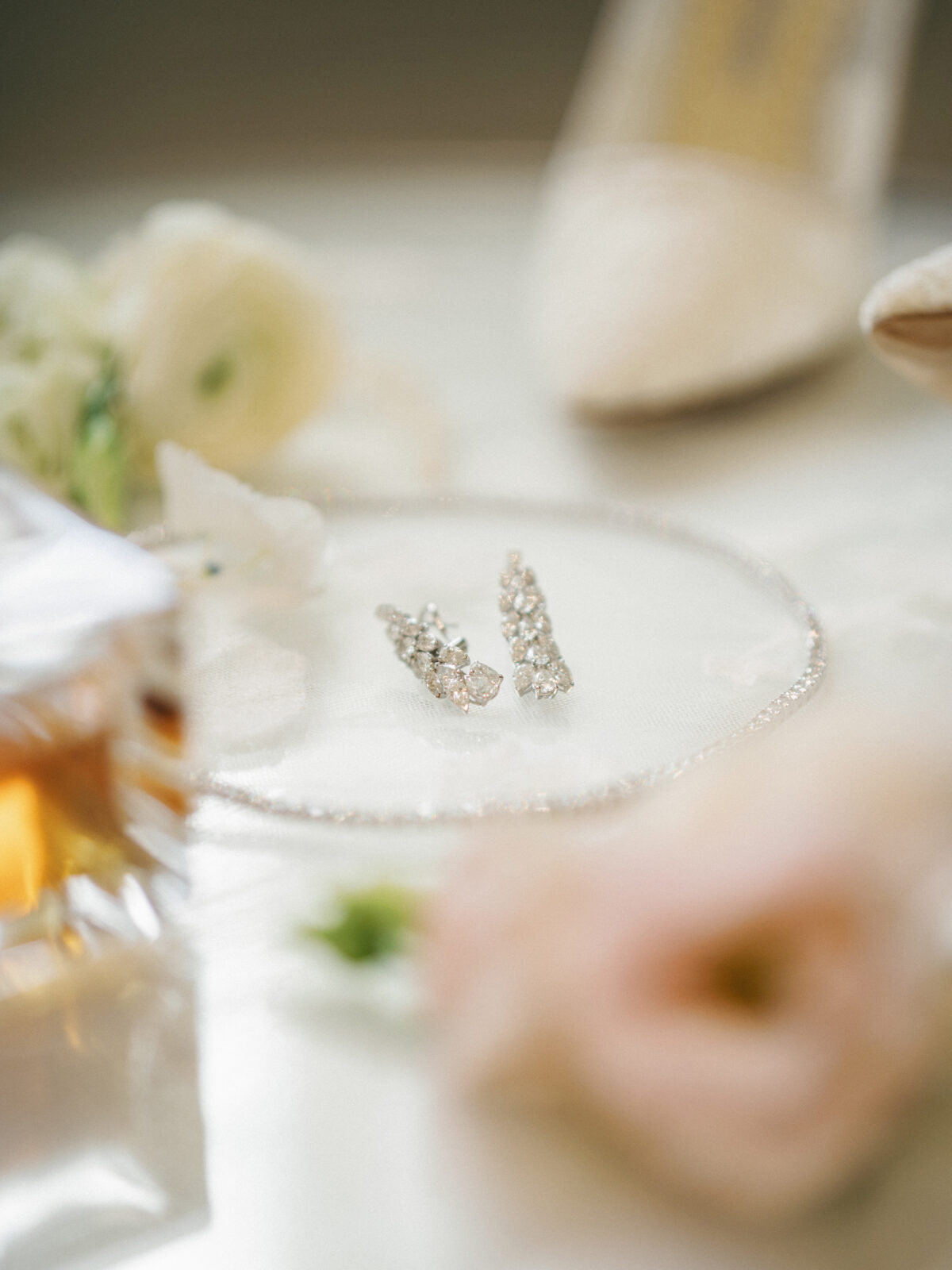 A pair of diamond earrings rests on a glass plate, surrounded by blurred flowers, a perfume bottle, and a high-heeled shoe in the background.