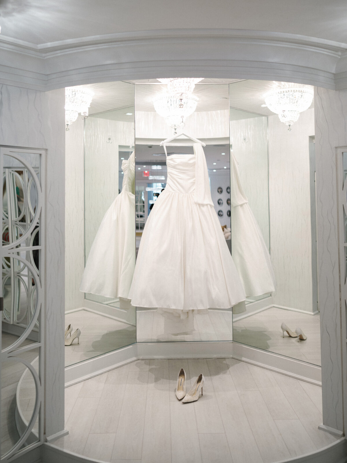 A white wedding dress is displayed on a hanger in front of mirrors under a chandelier, with a pair of white shoes on the floor below.