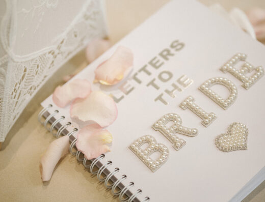 A spiral notebook labeled “Letters to the Bride” with pearl lettering, decorated with scattered pink rose petals, rests on a table next to a lace item.