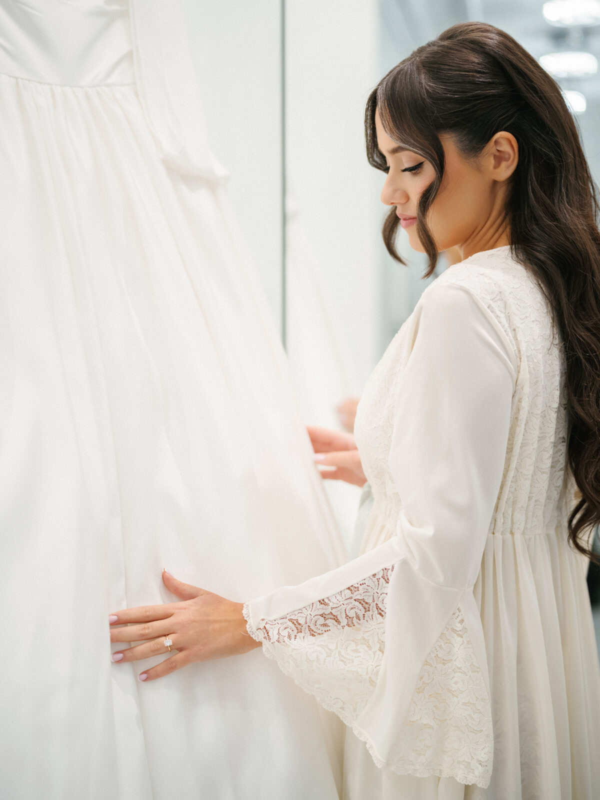 A woman with long dark hair gently touches a white wedding dress on display, wearing a white lace robe and looking at the gown.