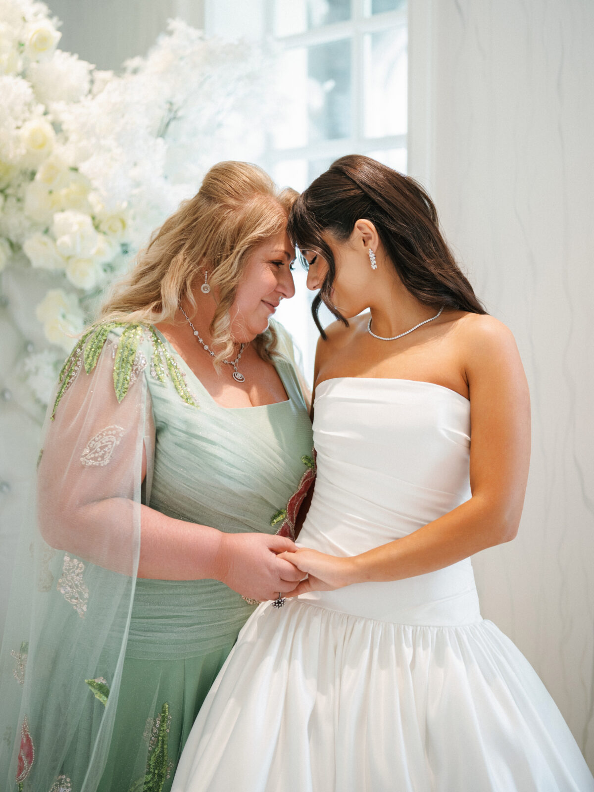 Two women in formal dresses stand close together, holding hands and touching foreheads, with white flowers and a window in the background.