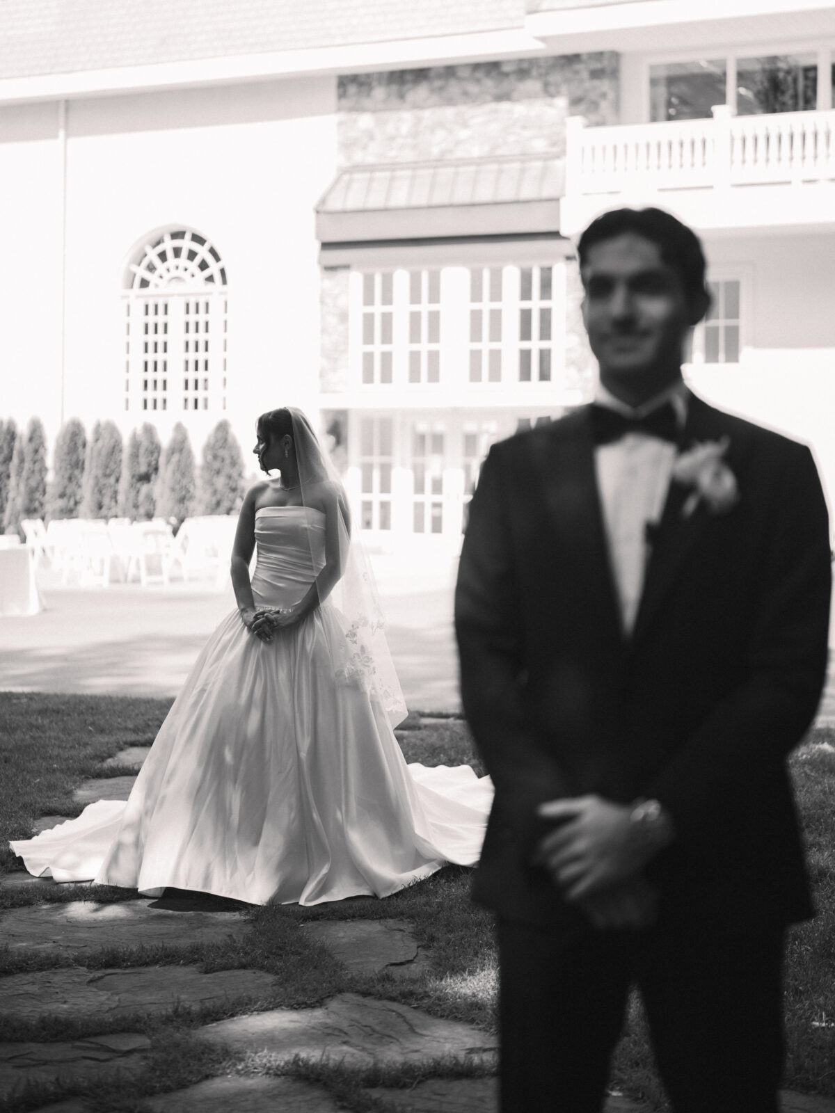 A bride in a wedding dress stands in the background while a groom in a suit stands out of focus in the foreground at an outdoor venue.