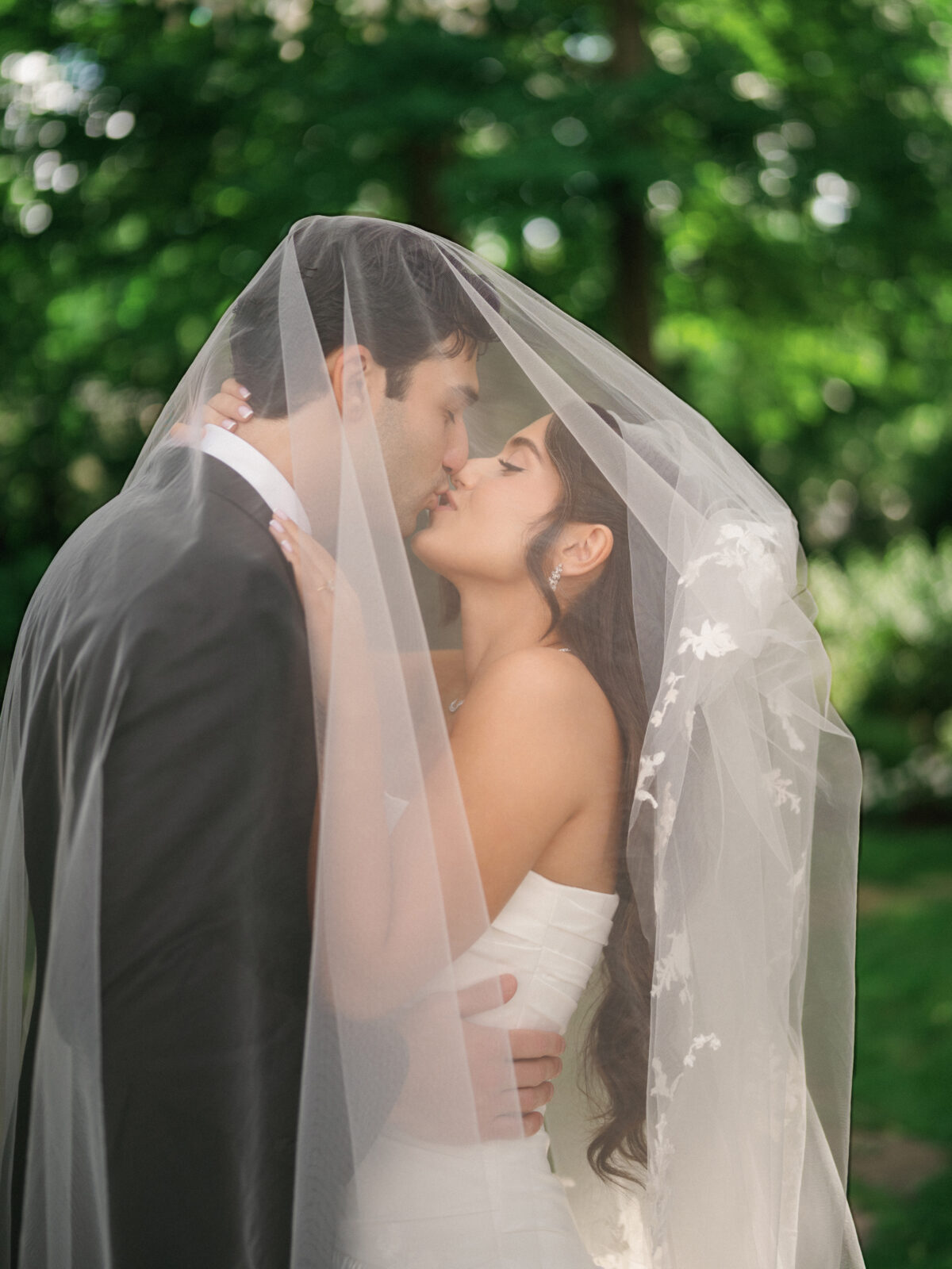 A bride and groom kiss under a sheer wedding veil outdoors, surrounded by green trees in soft, natural light.