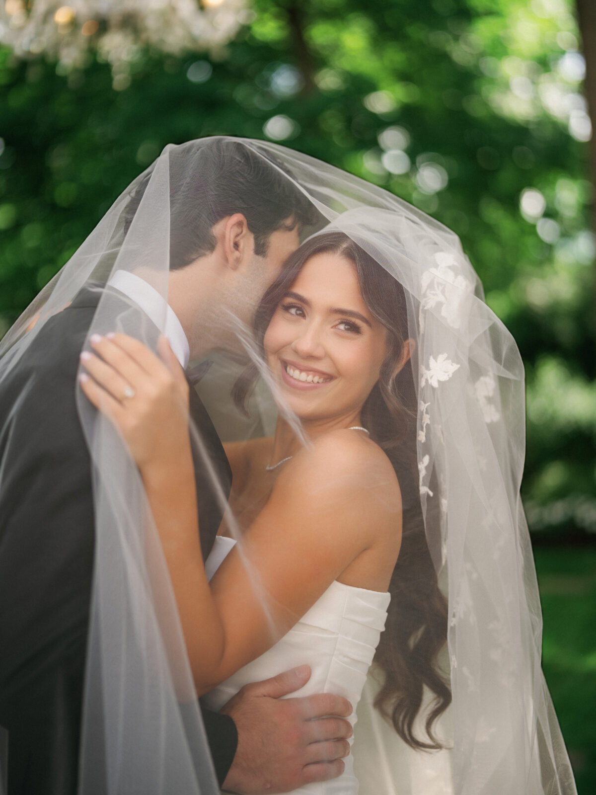 A bride and groom embrace under a sheer veil outdoors, with green foliage in the background. The bride smiles while looking away from the groom.