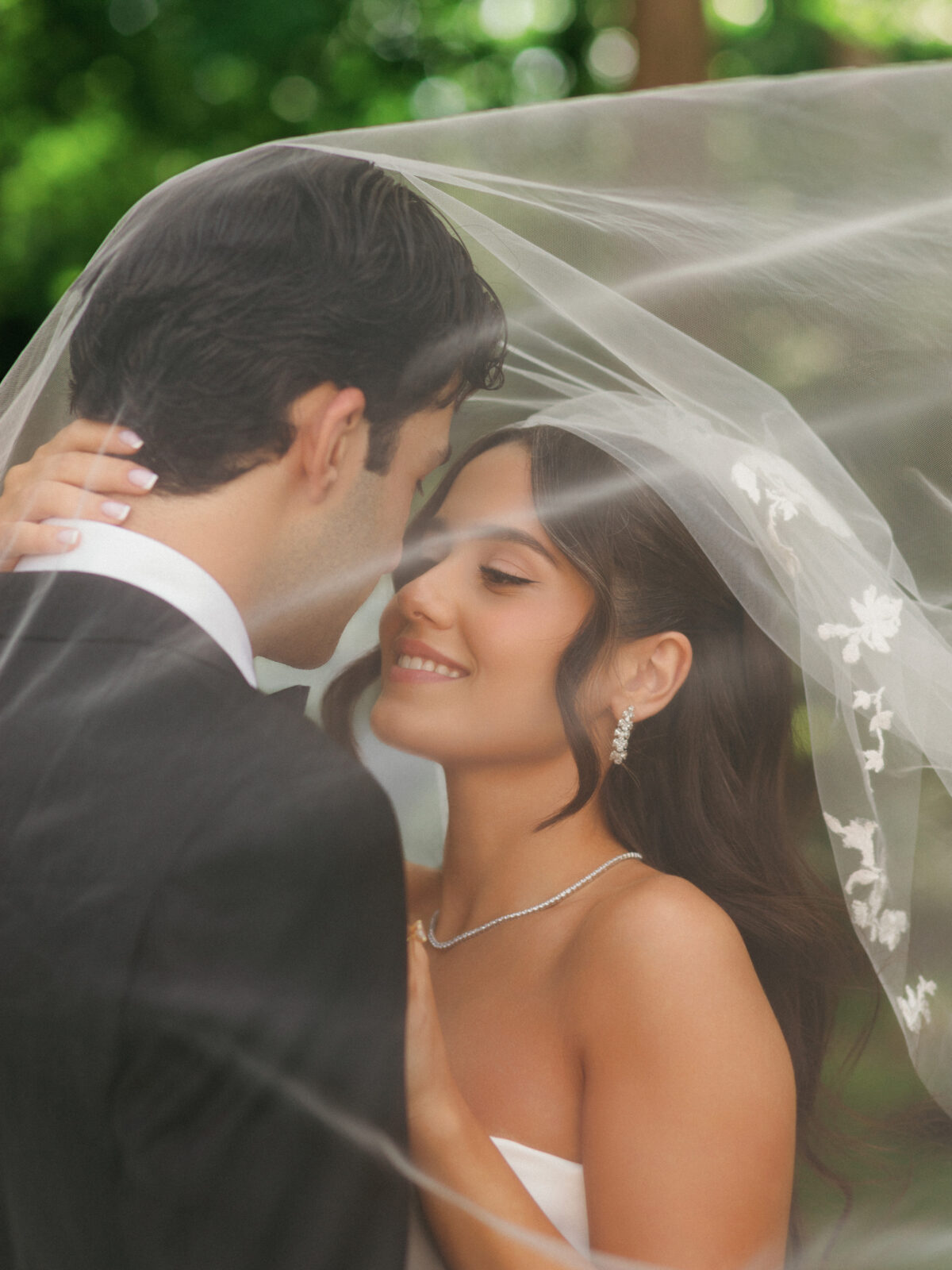 A bride and groom stand close together under a bridal veil outdoors, gazing at each other and smiling, with greenery in the background.