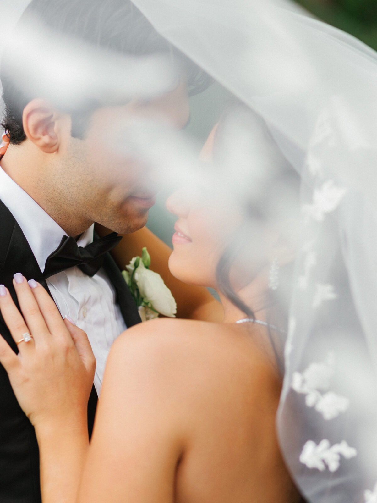 A bride and groom stand close together under a sheer veil, with her hand on his chest showing a ring and his boutonniere visible. Their faces are partially obscured by the veil.