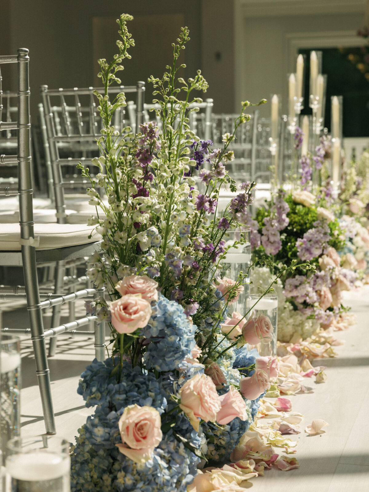 Rows of silver chairs line an aisle decorated with pastel flower arrangements, including roses and hydrangeas, with scattered flower petals on the floor.