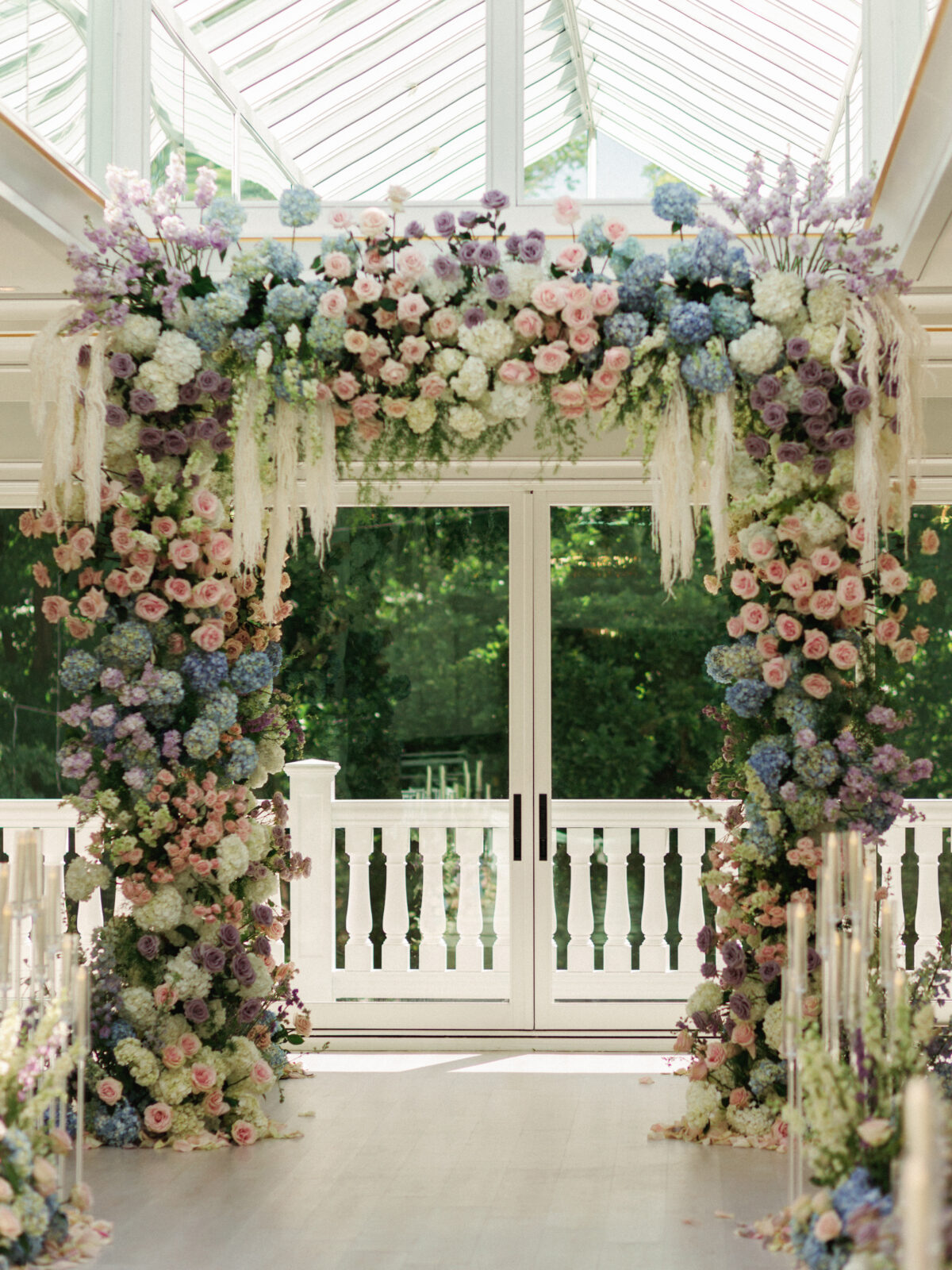 A floral archway with pink, purple, blue, and white flowers stands indoors in front of glass doors and windows, with greenery visible outside.