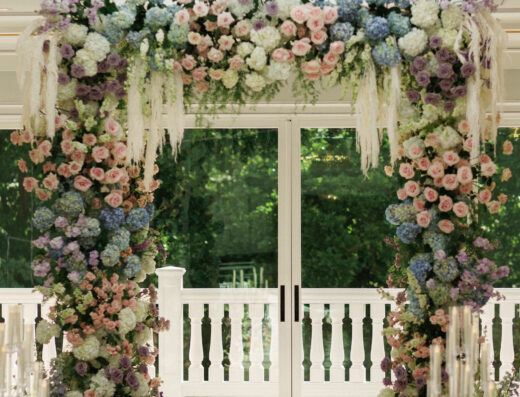 A floral archway with pink, purple, blue, and white flowers stands indoors in front of glass doors and windows, with greenery visible outside.