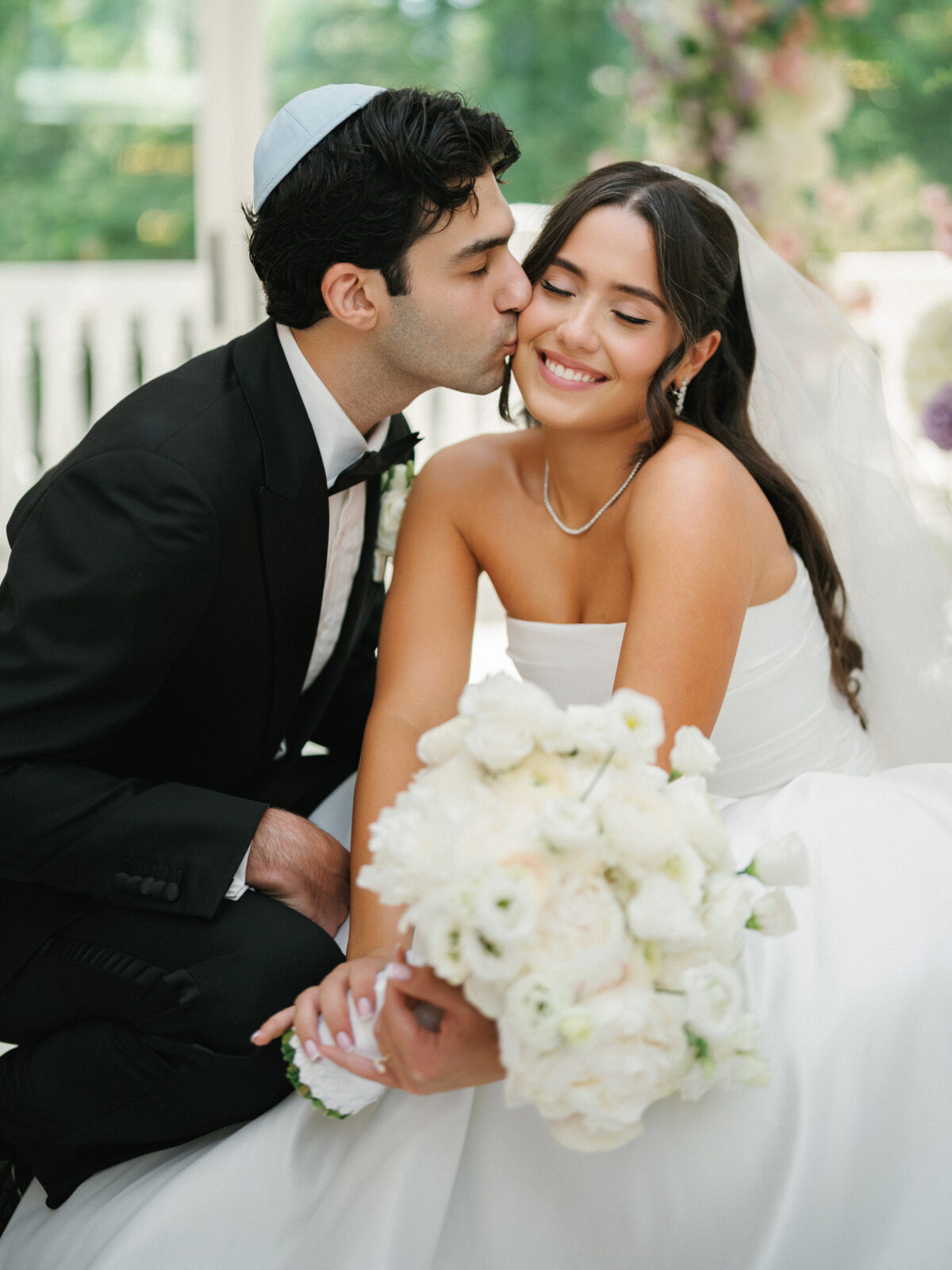 A groom in a black suit and kippah kisses the smiling bride on the cheek as she holds a bouquet of white flowers.