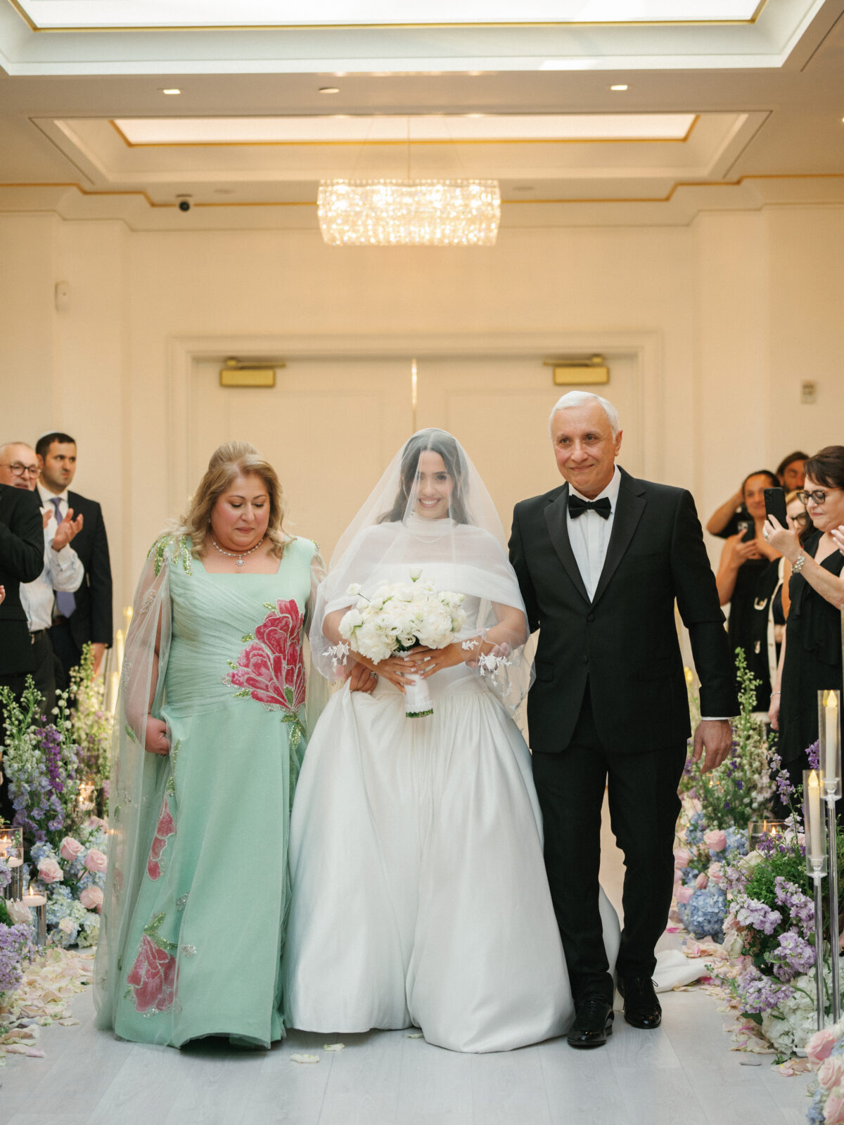 A bride in a white gown and veil walks down the aisle holding a bouquet, accompanied by two adults, surrounded by guests and floral arrangements.