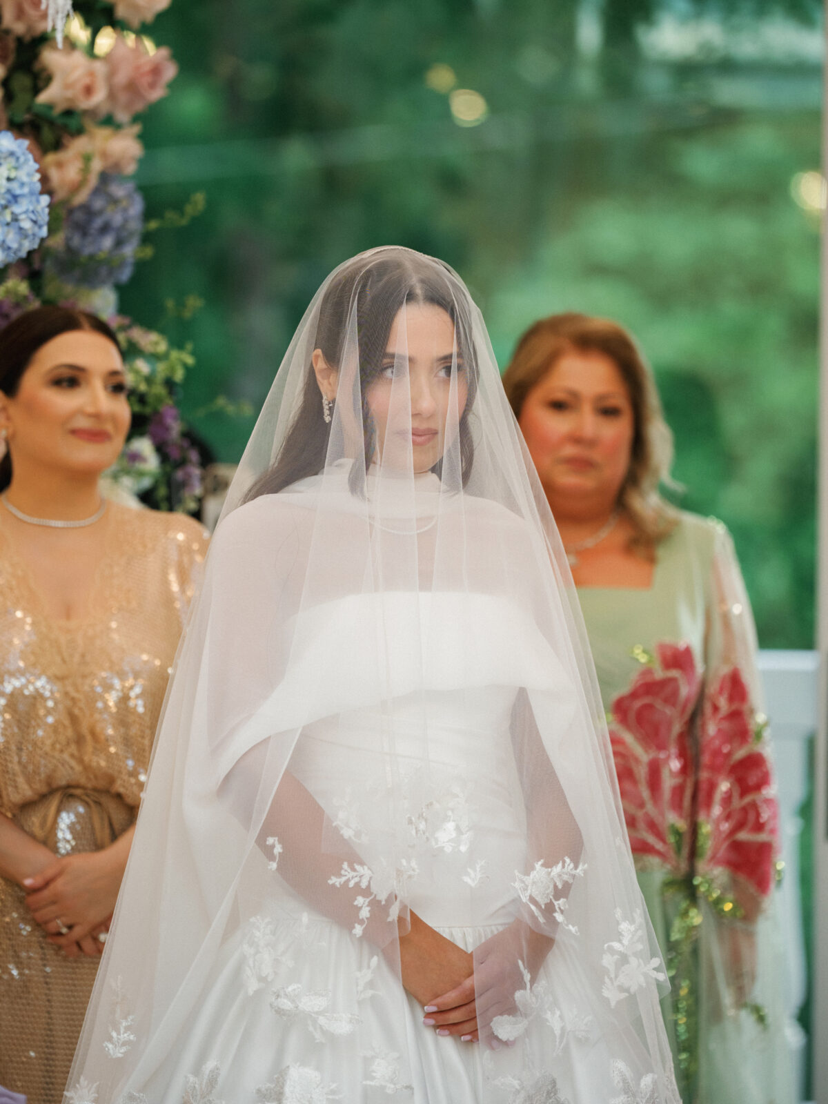 A bride in a white dress and veil stands with her hands clasped, flanked by two women in formal attire; floral decorations are visible in the background.